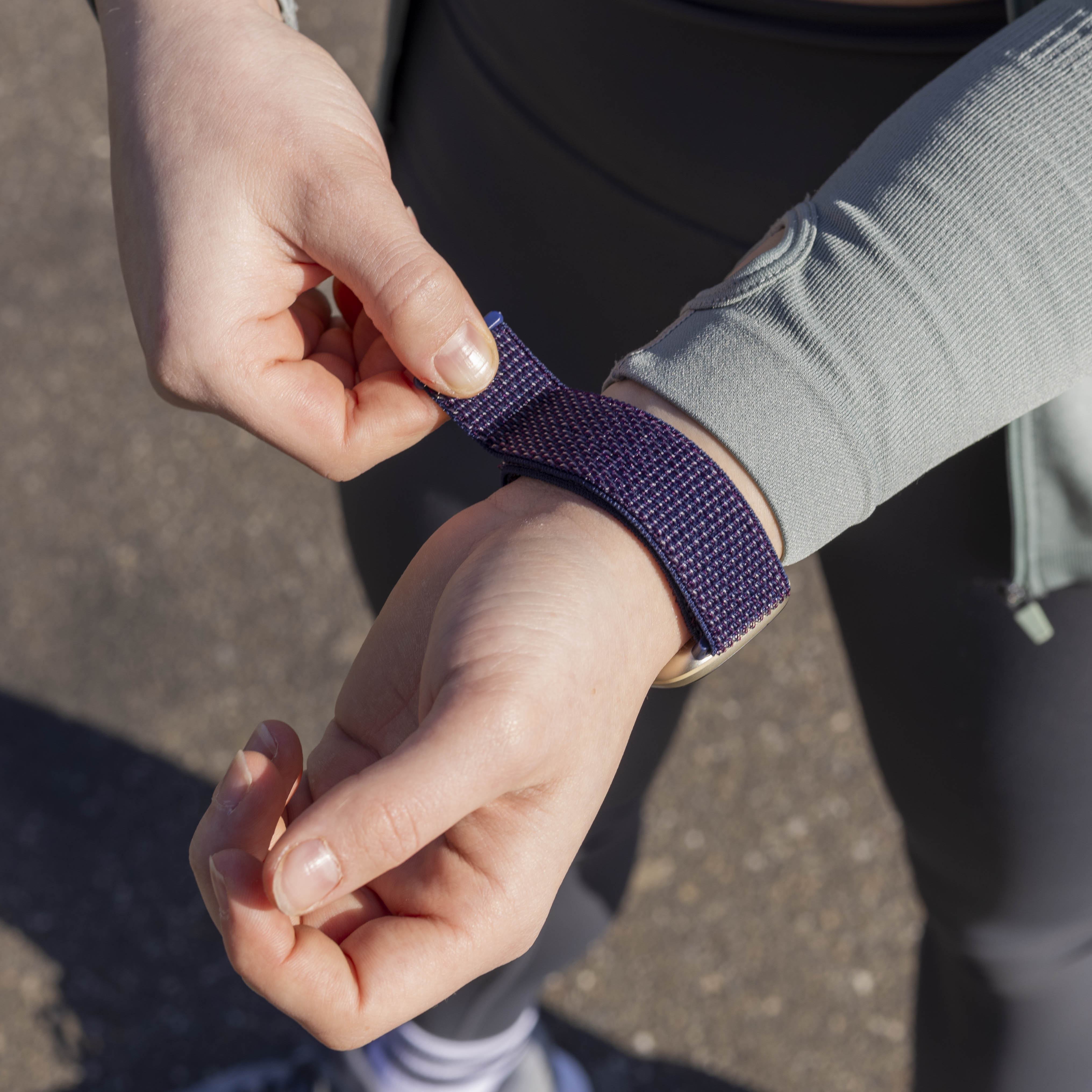 Eine Person passt ein lila Uhrenarmband an ihrem Handgelenk an. Graue Kleidung und Asphalt sind sichtbar.