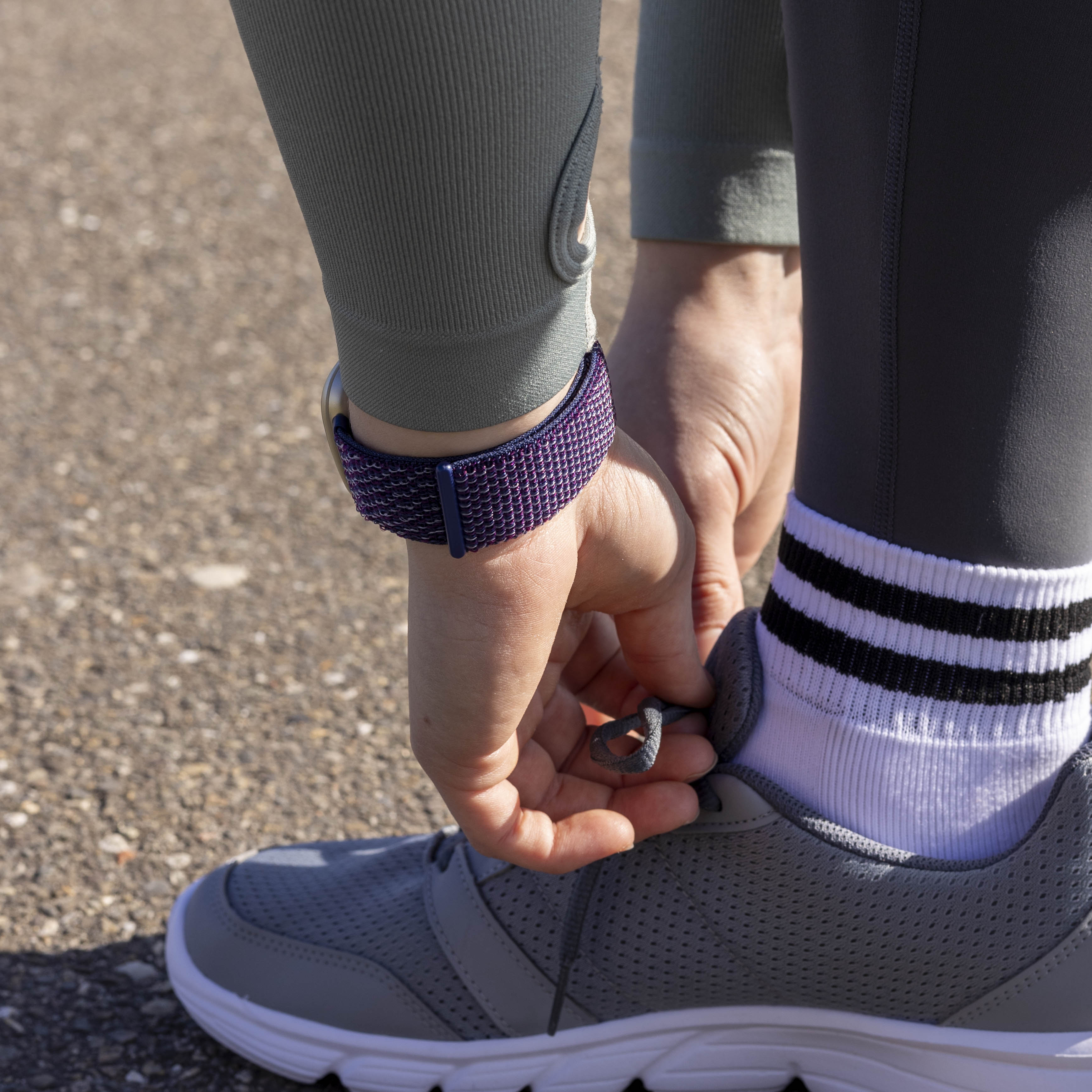 Eine Person bindet einen grauen Laufschuh. Sie trägt eine lila Uhr, graue Leggings und weiße Socken mit schwarzen Streifen.