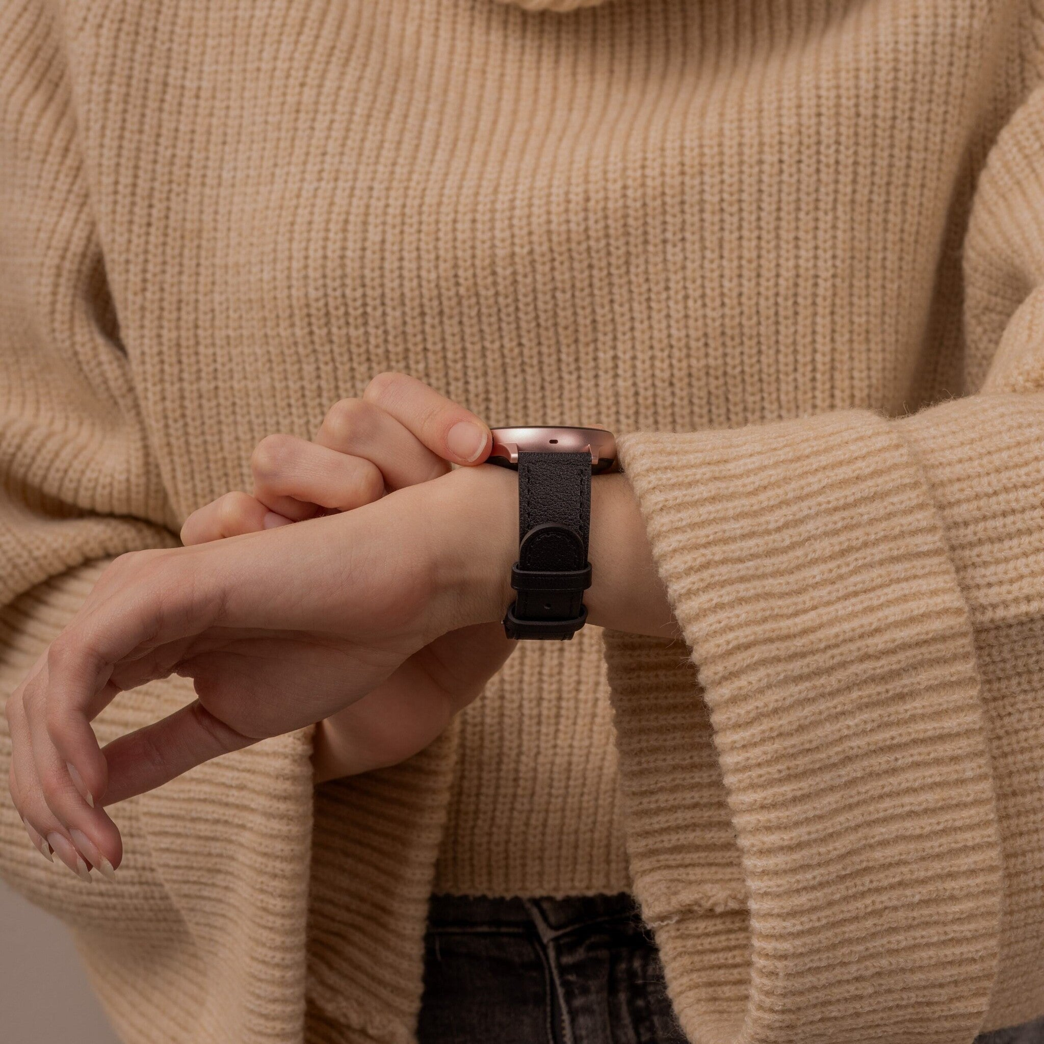 Eine Person in einem beigefarbenen Pullover schaut auf ihre Uhr. Die Uhr ist roségold mit einem schwarzen Lederarmband.