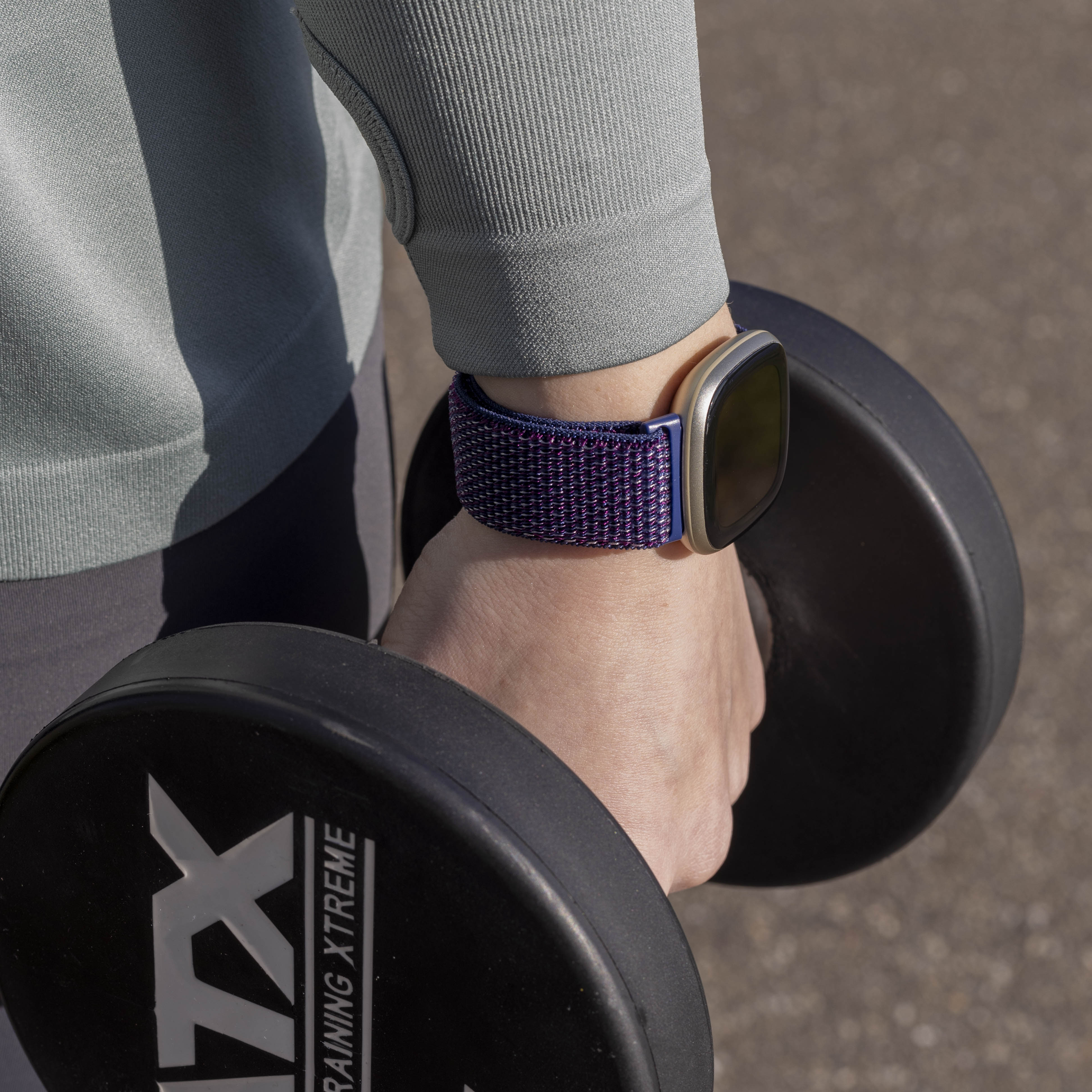 Eine Person hält eine Hantel. Sie trägt eine Uhr mit blau-lila Armband und ein graues Langarmshirt.