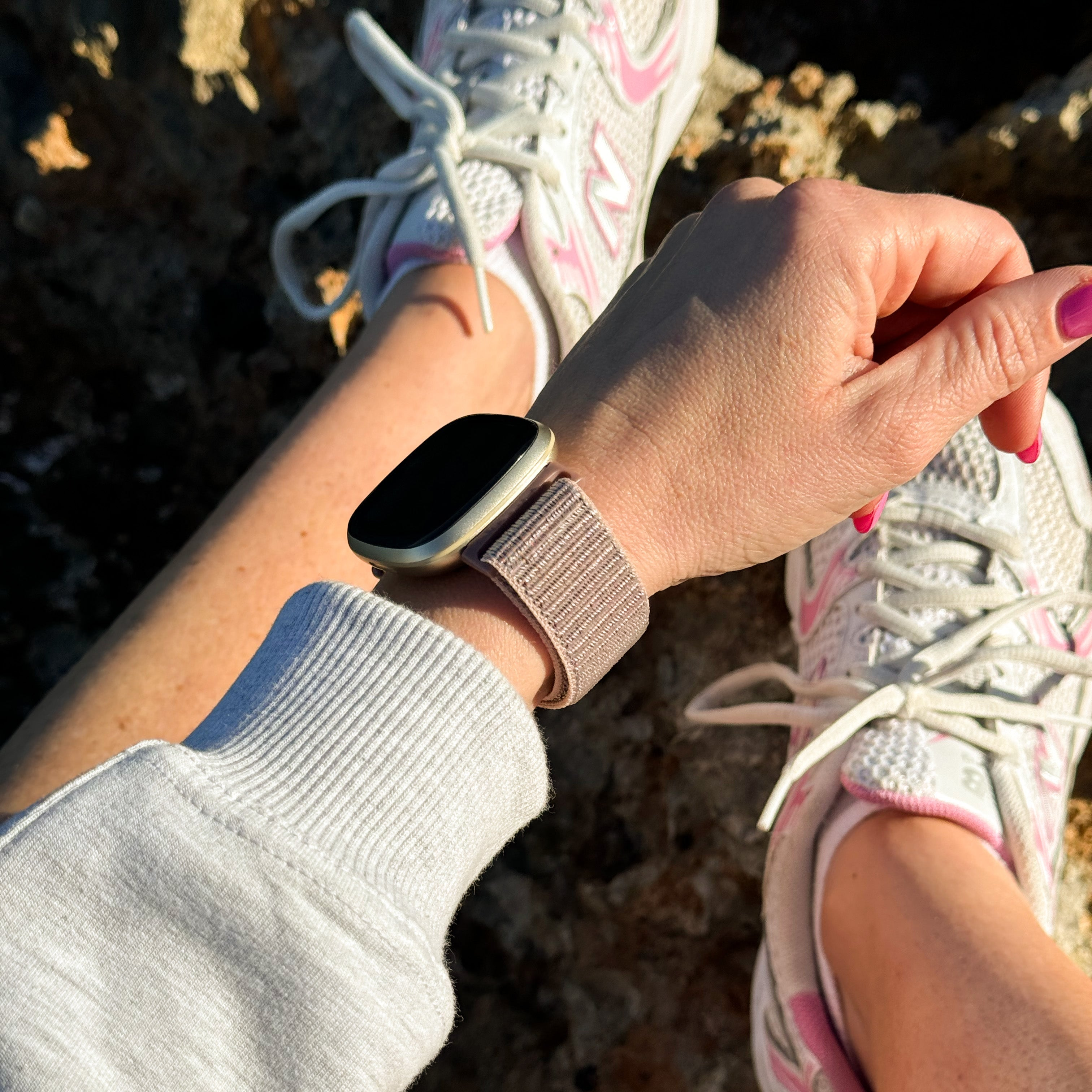 Der Arm einer Person mit einer Uhr ist sichtbar. Die Person trägt weiße Turnschuhe mit rosa Akzenten. Die Nägel sind rosa lackiert.