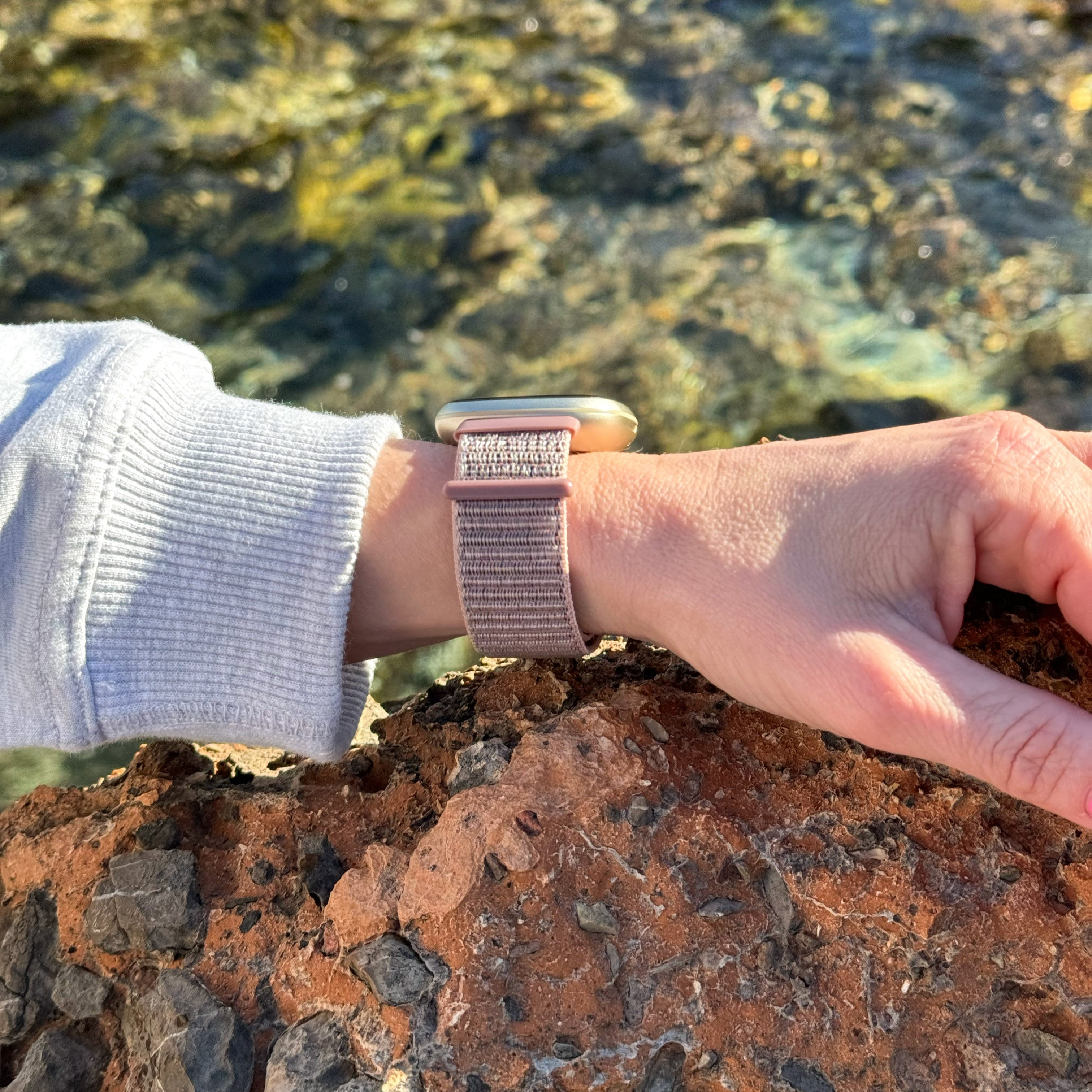 Handgelenk mit Smartwatch, Händen und einem Felsen. Die Uhr hat ein rosa Armband. Der Hintergrund ist verschwommen.