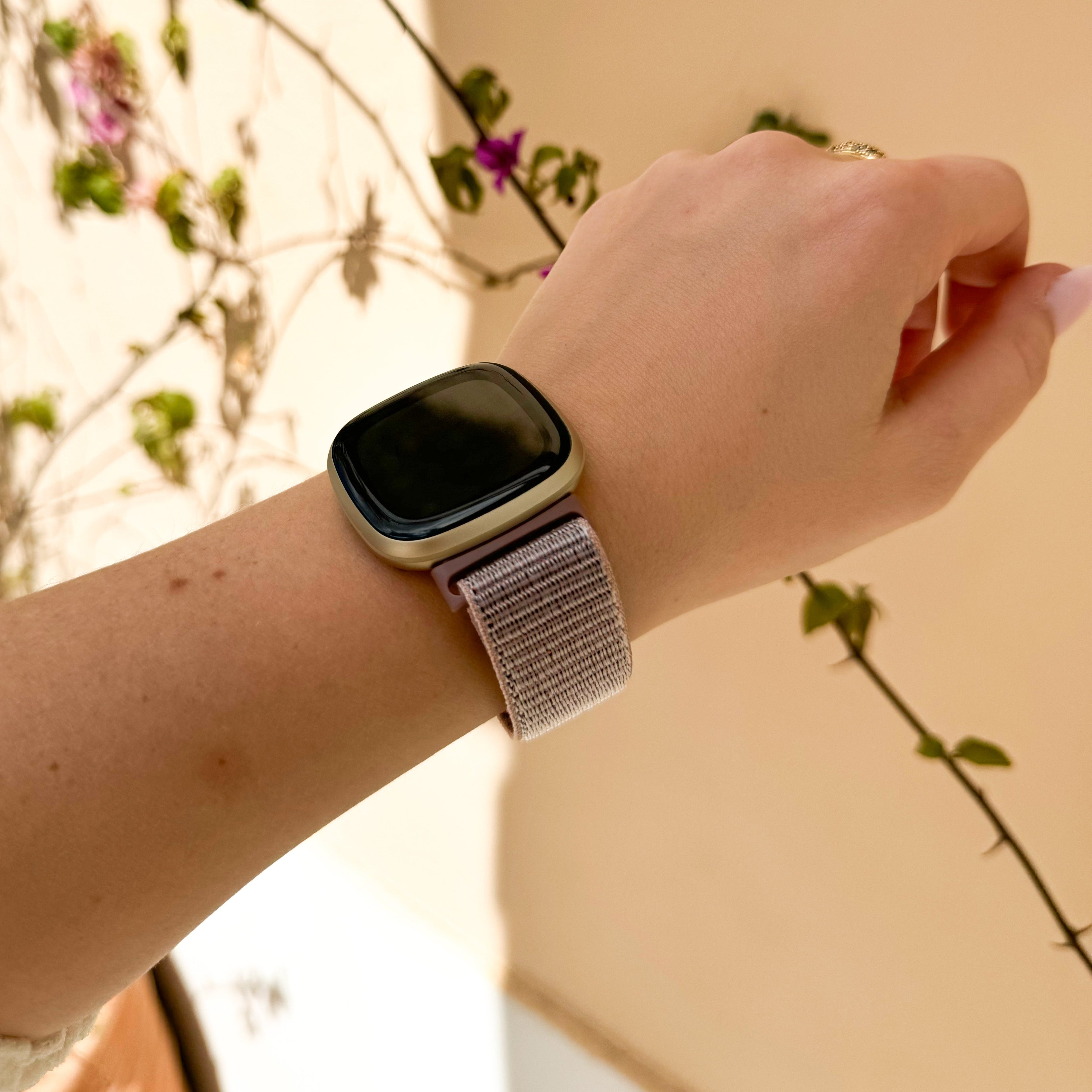 Ein Arm mit Smartwatch, Ring und Pflanzenstängeln im Hintergrund. Neutraler Hintergrund.