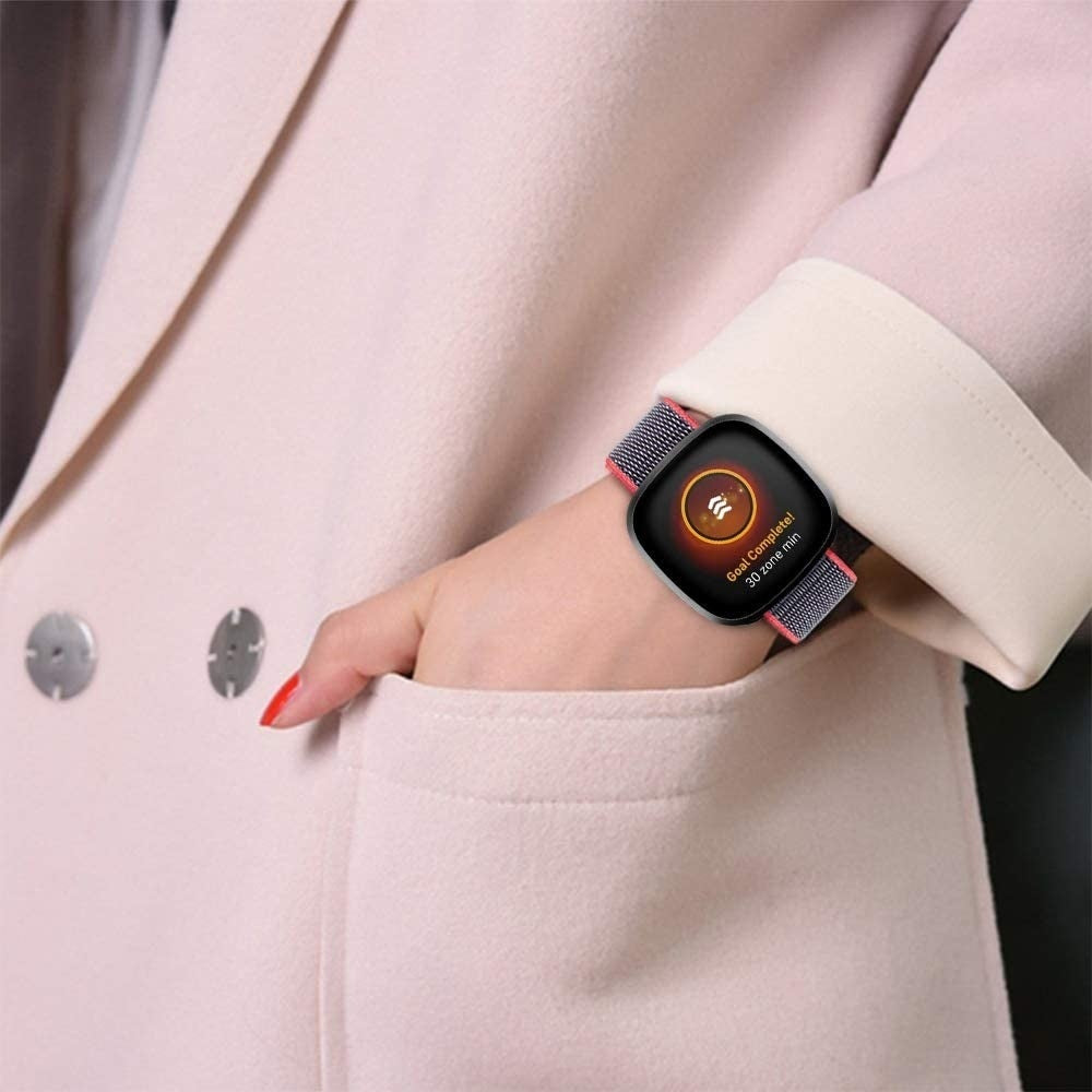 Das Handgelenk einer Frau mit einer Smartwatch, die 'Ziel erreicht' anzeigt. Die Uhr hat ein graues und rosa Armband. Die Frau trägt einen rosa Blazer.