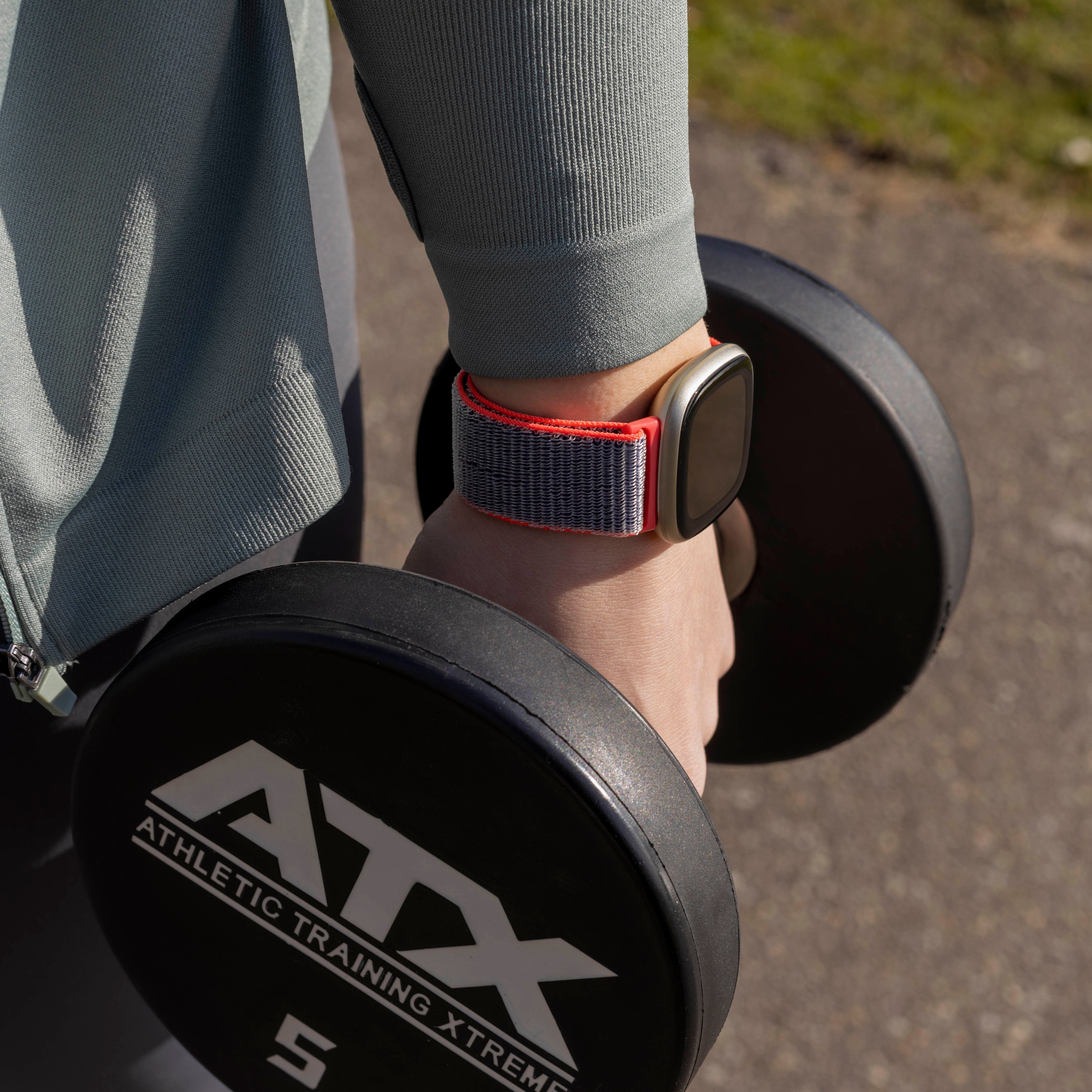 Eine Person hält eine Hantel, trägt eine Fitnessuhr mit grau-rotem Armband und eine graue Jacke.