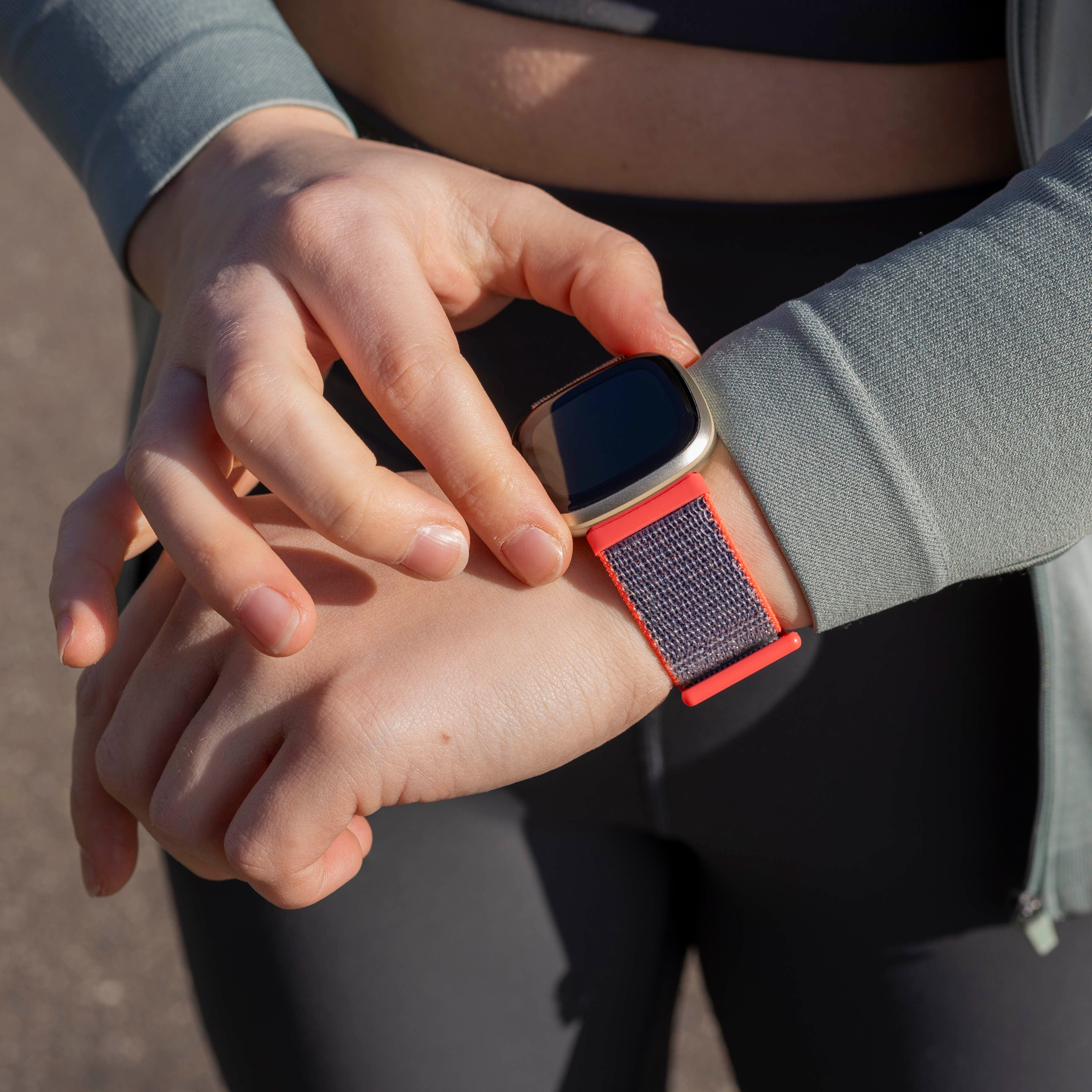 Person passt Smartwatch an. Die Uhr ist goldfarben mit rotem Armband. Person trägt graue Jacke und schwarze Leggings.