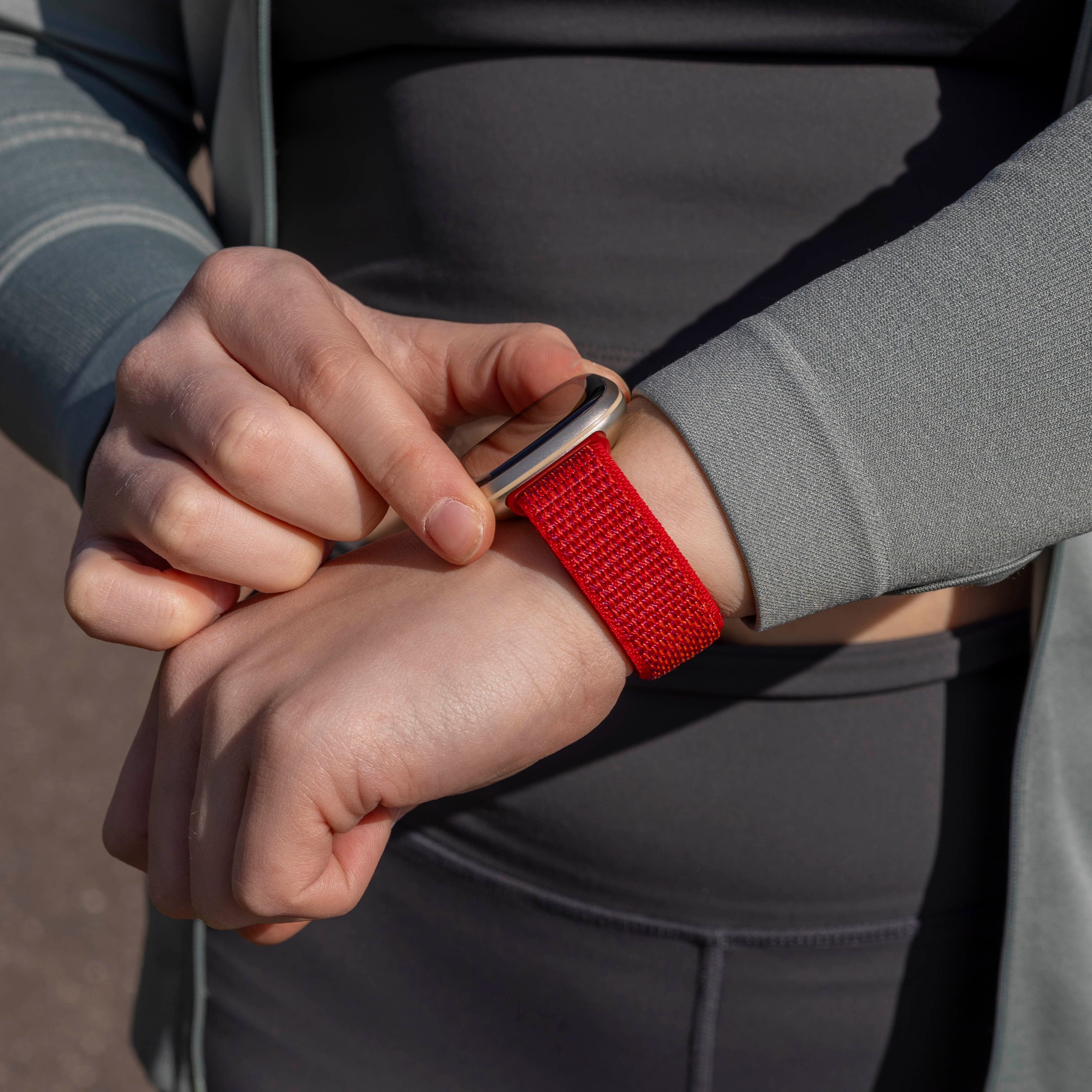 Das Handgelenk einer Person mit rotem Uhrenarmband. Die Hände der Person sind sichtbar, sie trägt eine graue Jacke und ein Hemd.