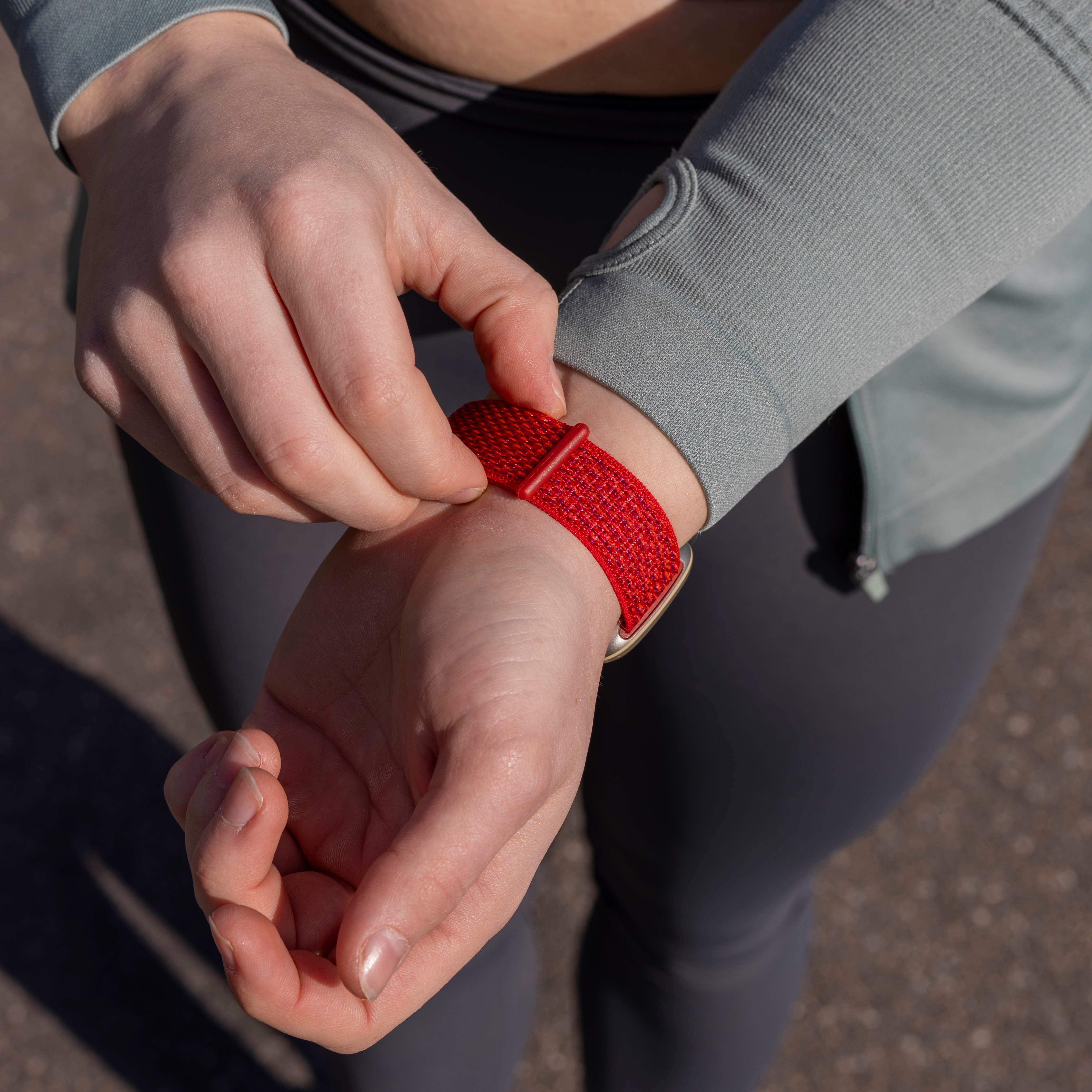 Hände einer Person, die ein rotes Armband einstellen. Sie trägt einen grauen Langarm und dunkelgraue Leggings. Der Hintergrund ist unscharf.