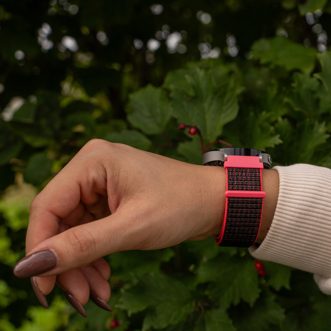 Eine Person trägt eine Smartwatch am Handgelenk. Das Armband ist rosa und schwarz. Im Hintergrund grünes Laub.