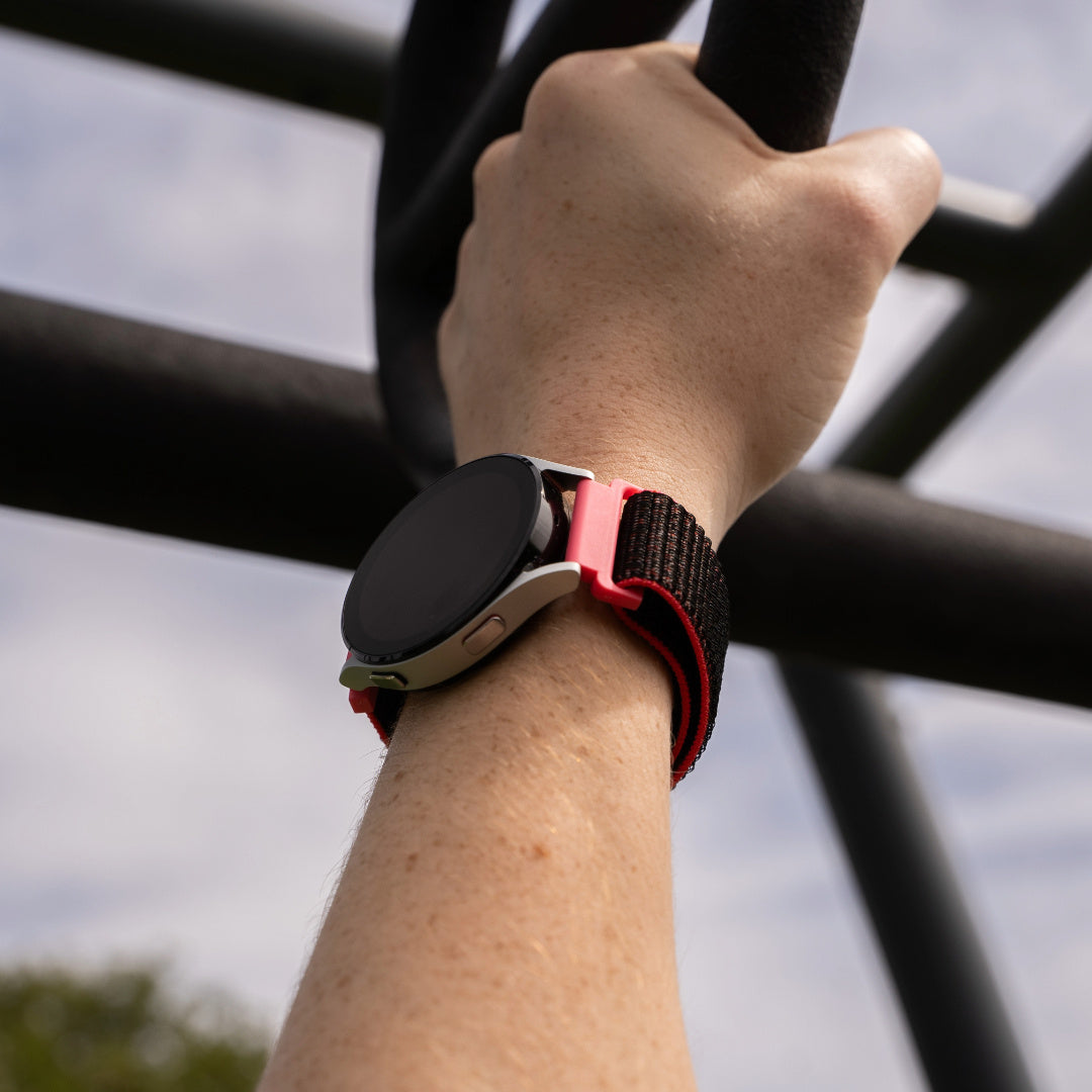 Ein Arm hält eine schwarze Stange. Eine Smartwatch mit schwarzem und rotem Armband befindet sich am Handgelenk. Der Hintergrund ist ein bewölkter Himmel.