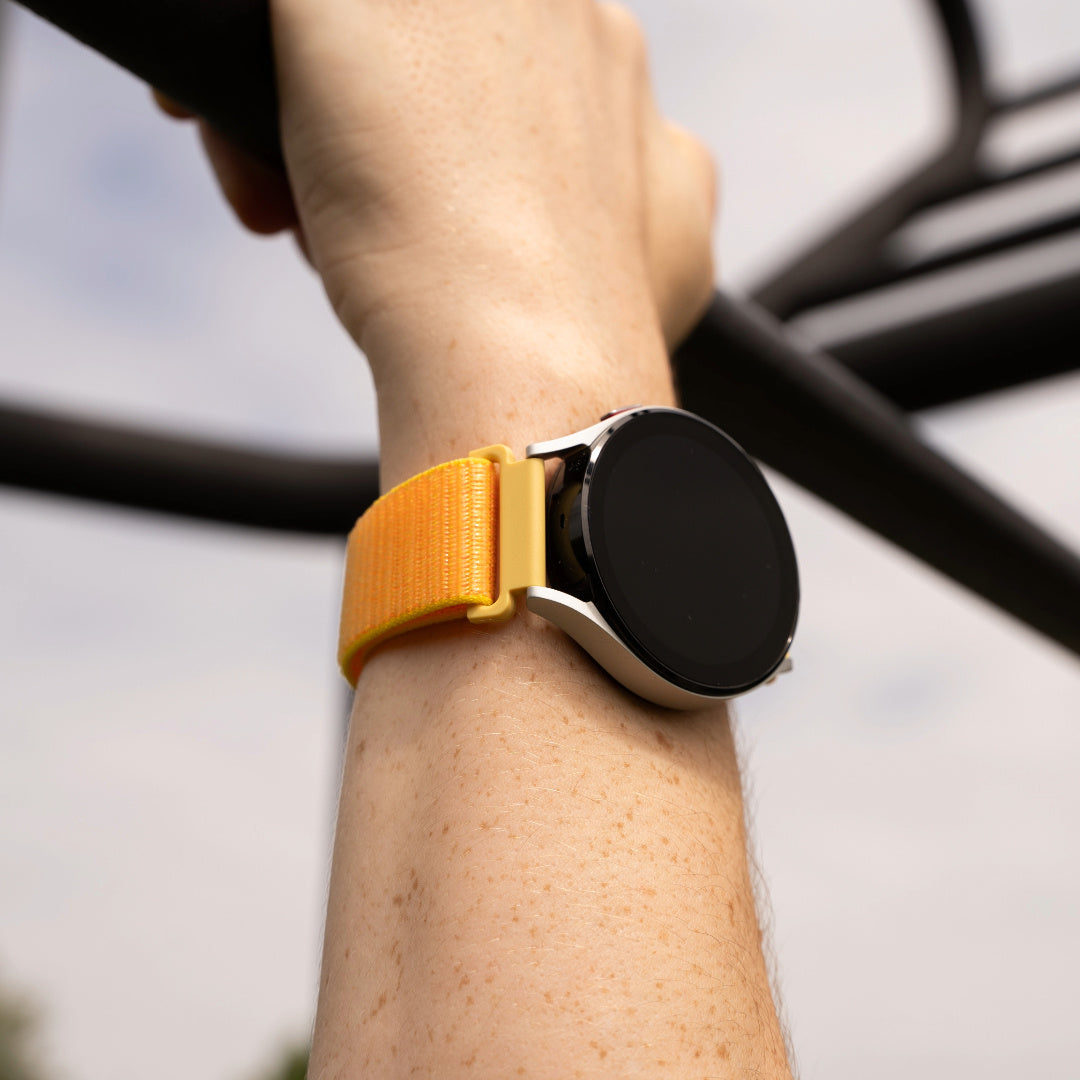 Ein Handgelenk hält eine Smartwatch mit orangefarbenem Armband. Die Uhr ist schwarz. Der Arm hält eine schwarze Stange mit unscharfem Hintergrund.