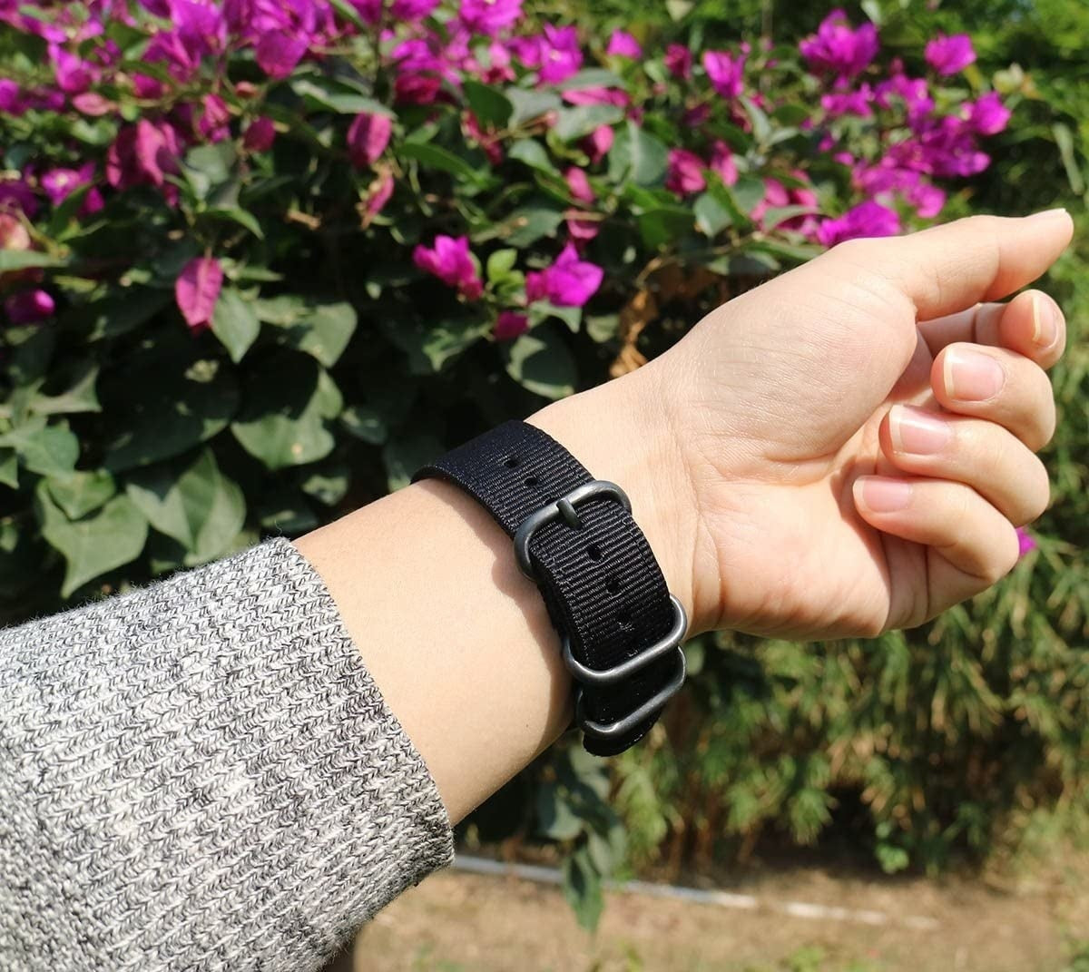 Ein Handgelenk mit schwarzem Uhrenarmband wird vor einem Hintergrund aus lila Blumen und grünen Blättern gezeigt.