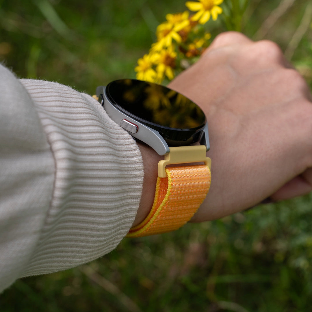 Handgelenk mit Smartwatch, gelb-orangefarbenem Armband und grünem Hintergrund mit gelben Blumen.
