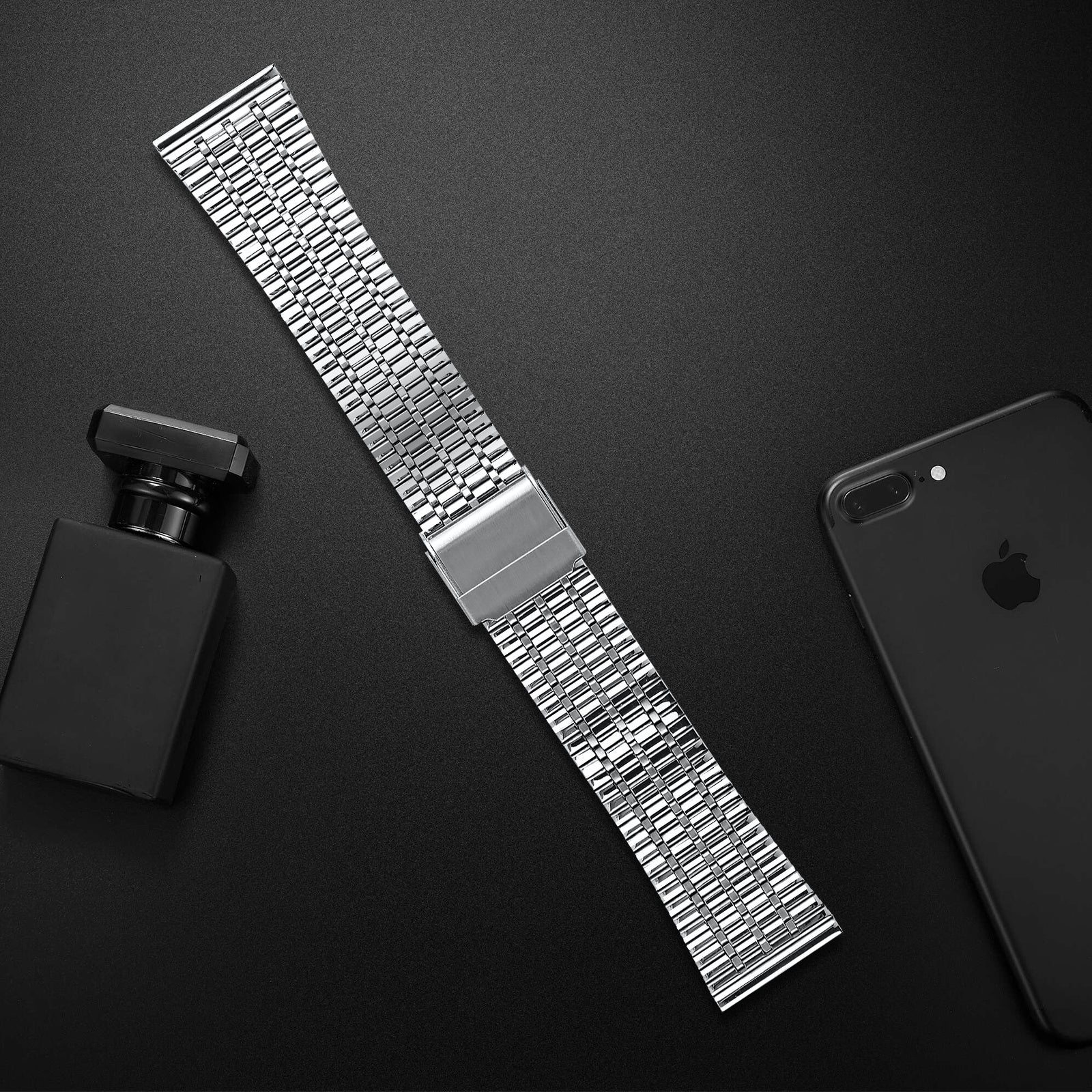 Ein silbernes Uhrenarmband, eine schwarze Parfümflasche und ein schwarzes iPhone sind auf einer schwarzen Oberfläche angeordnet.