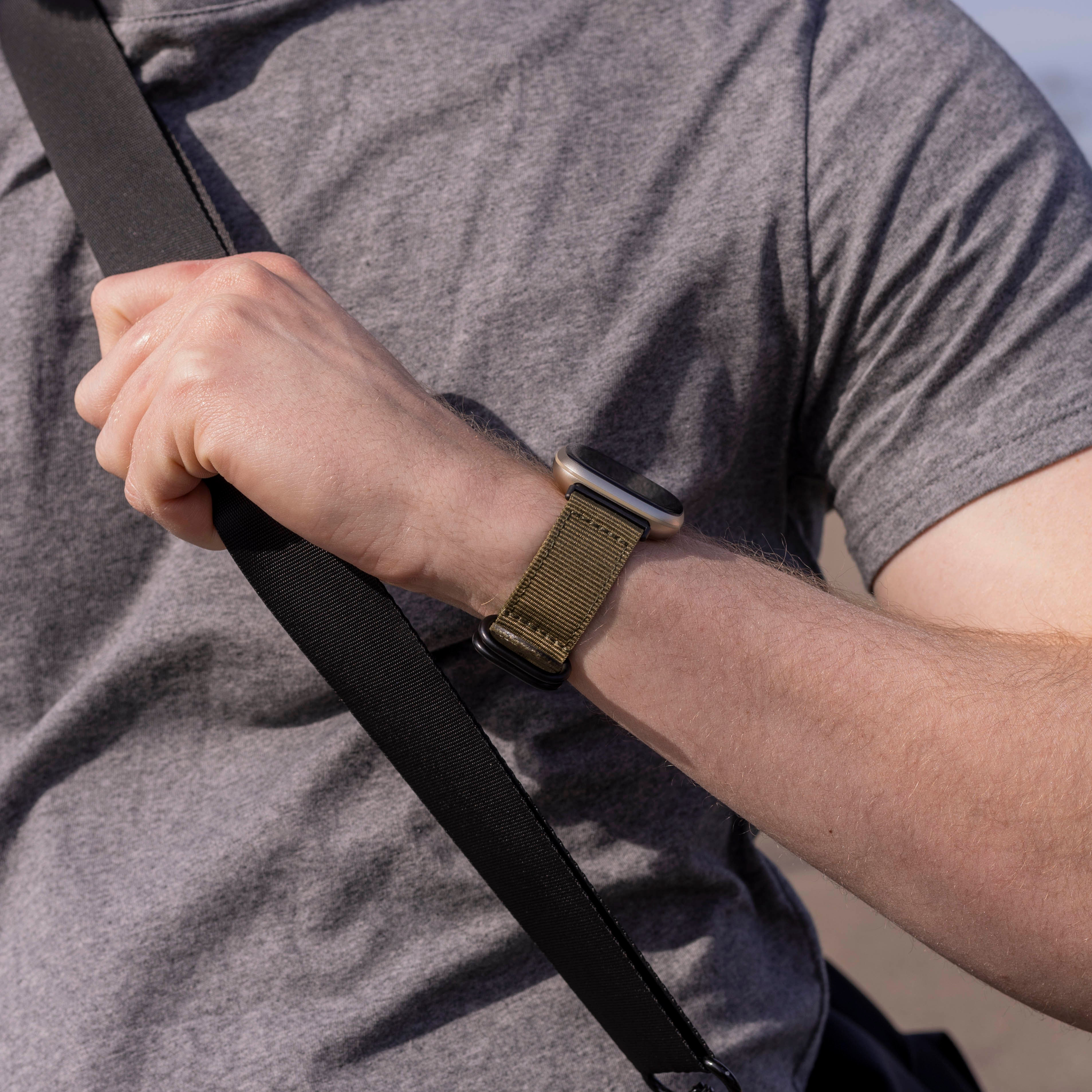 Eine Person trägt ein graues T-Shirt und eine Uhr mit olivgrünem Armband. Ein schwarzer Gurt ist ebenfalls sichtbar.