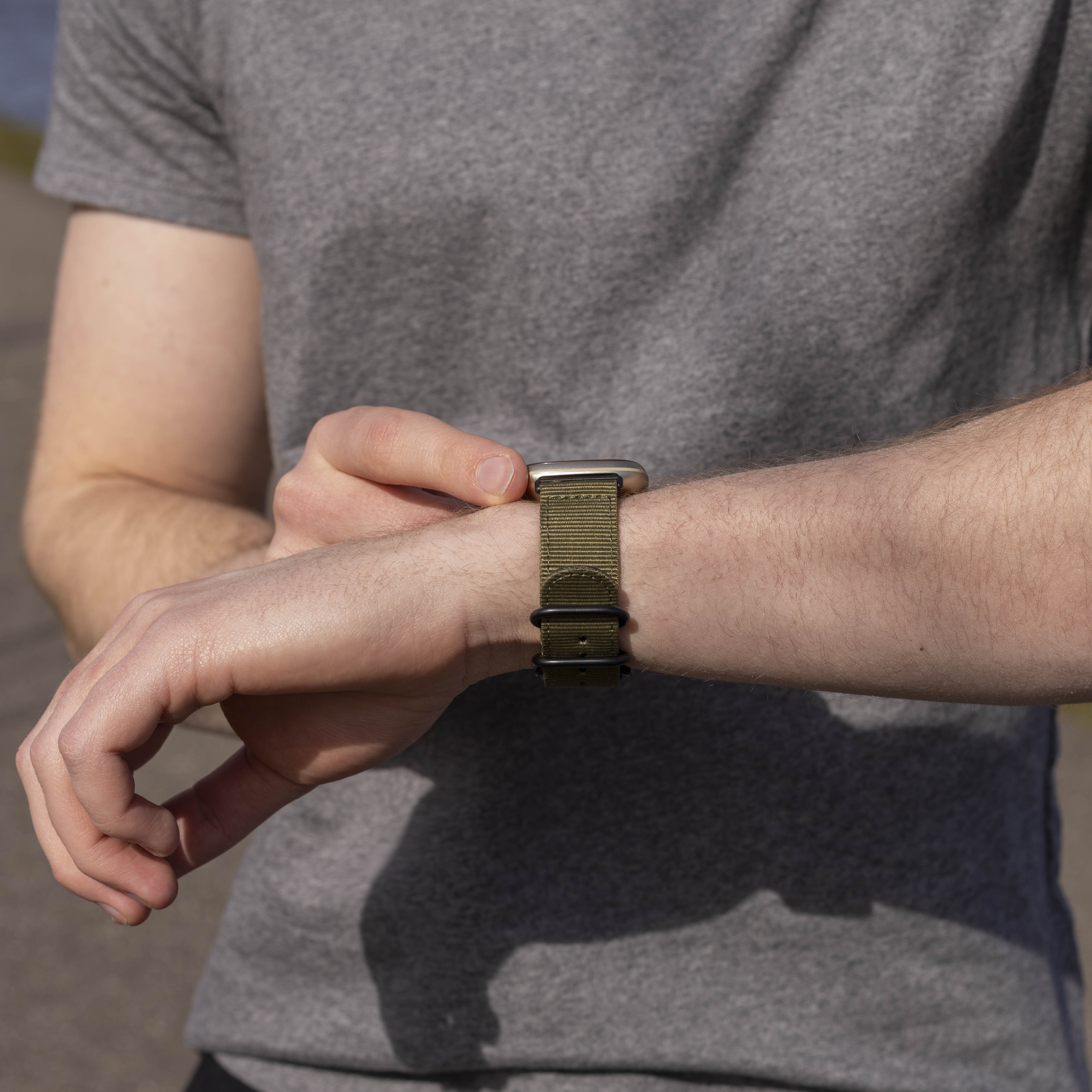 Eine Person in einem grauen Hemd schaut auf einer Smartwatch mit olivgrünem Armband auf die Uhrzeit.