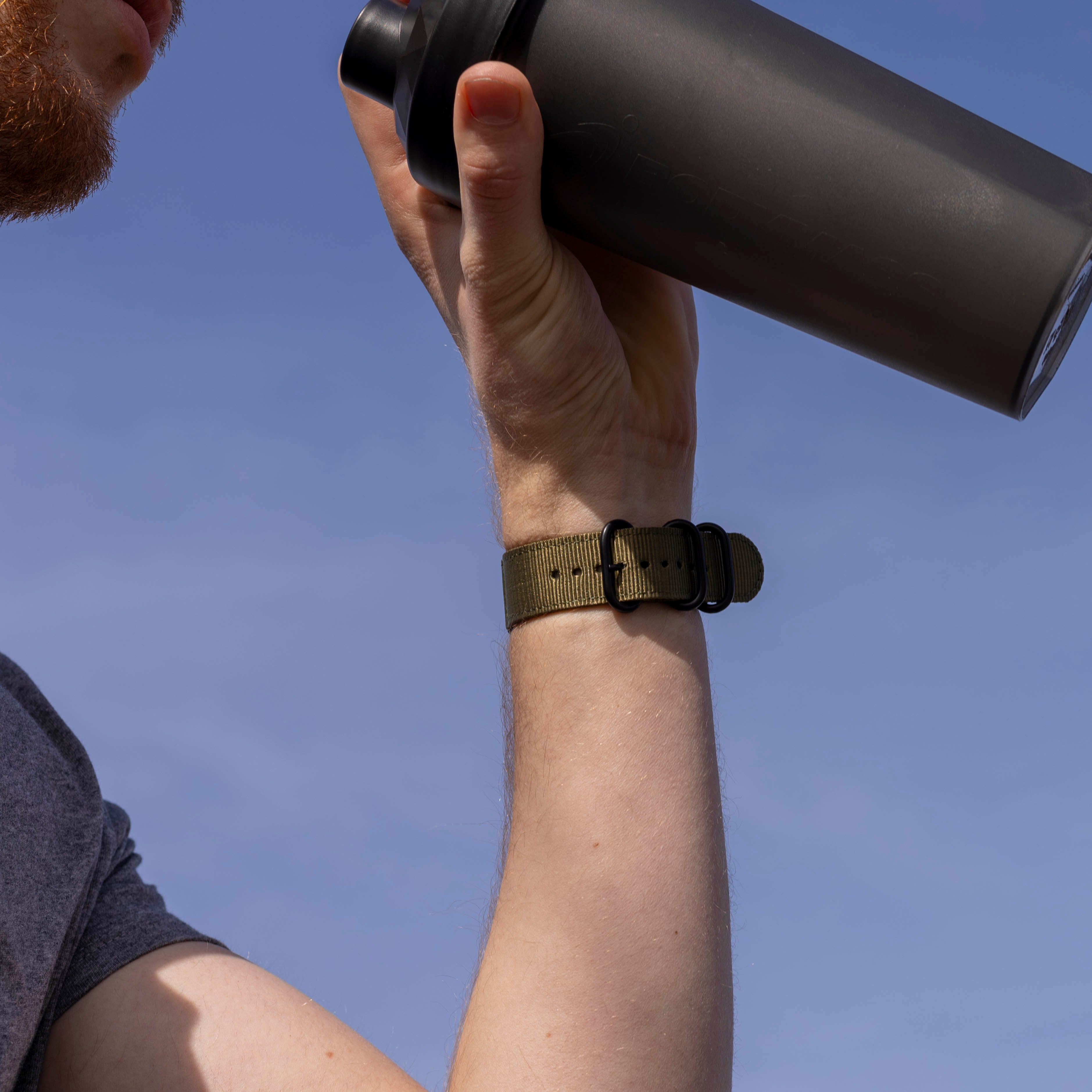 Person trinkt aus einem schwarzen Shaker. Arm mit olivfarbenem Armband sichtbar. Blauer Himmel im Hintergrund.