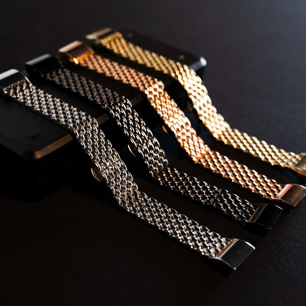 Vier Metall-Uhrenarmbänder in Silber, Schwarz, Roségold und Gold liegen auf einer schwarzen Oberfläche. Sie sind diagonal angeordnet.