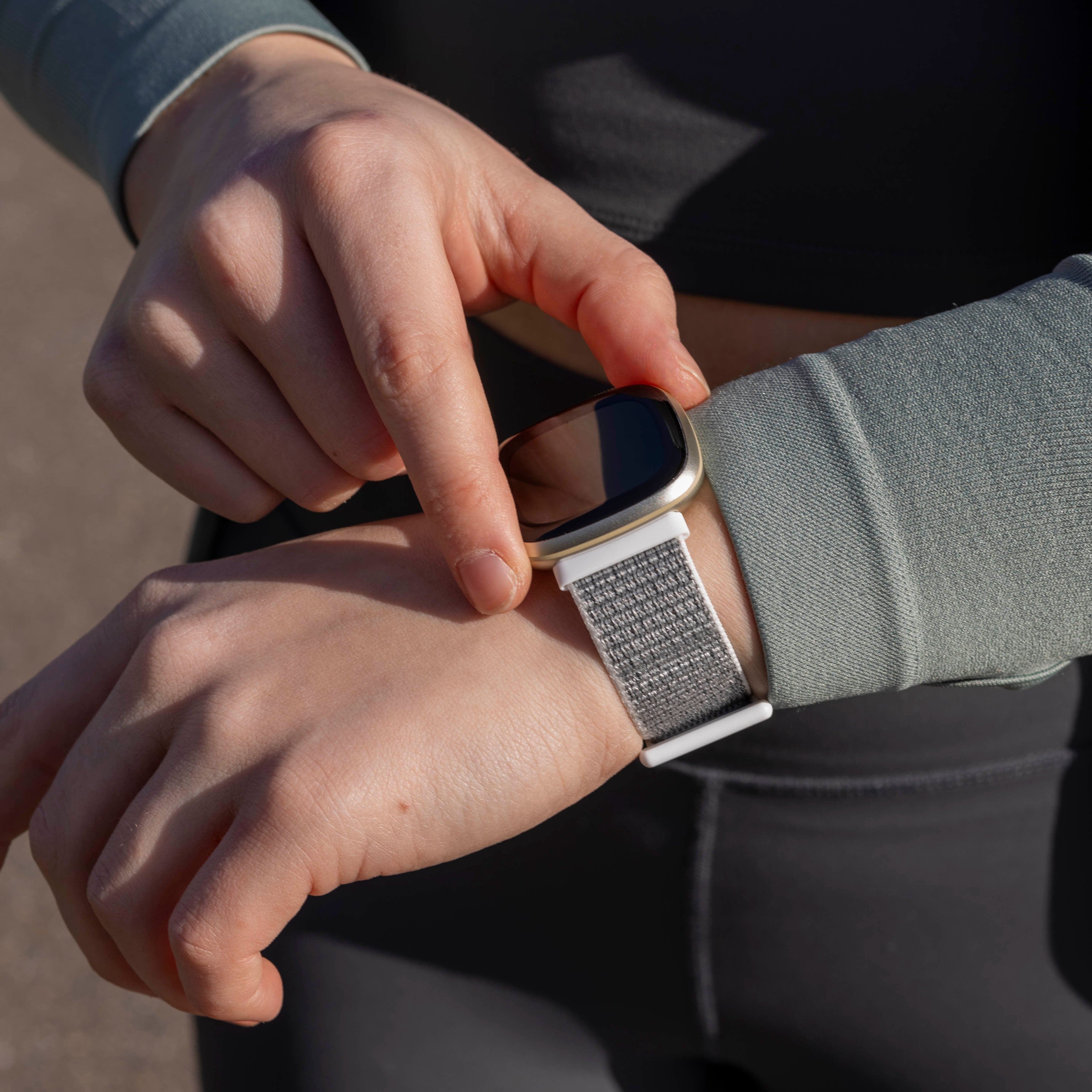 Hände einer Person, die eine Smartwatch mit goldenem Rahmen und grauem Armband am Handgelenk einstellt. Die Person trägt grüne Ärmel.