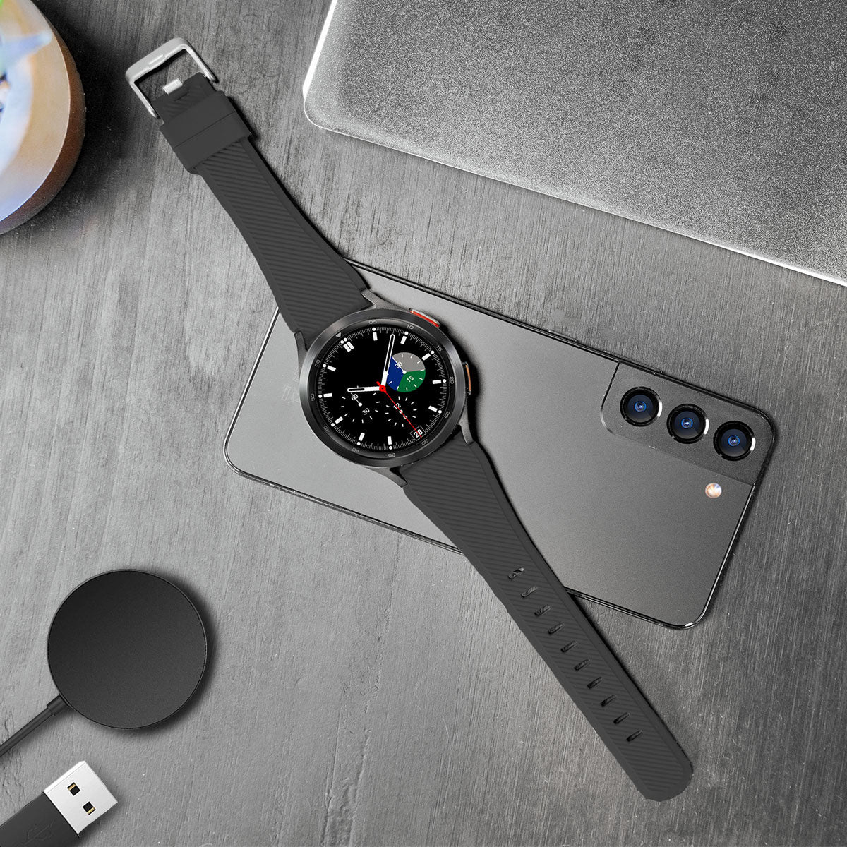 Schwarze Smartwatch auf einem Telefon. Ladegerät, Laptop und Stifthalter sind ebenfalls auf einer grauen Holzoberfläche zu sehen.