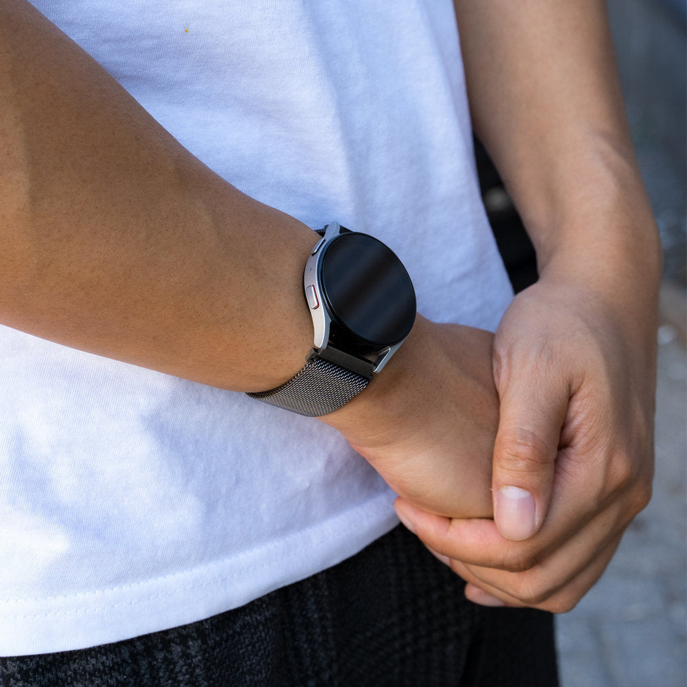 Eine Person trägt eine Smartwatch mit schwarzem Bildschirm und Metallarmband. Die Person trägt ein weißes T-Shirt. Der Hintergrund ist unscharf.