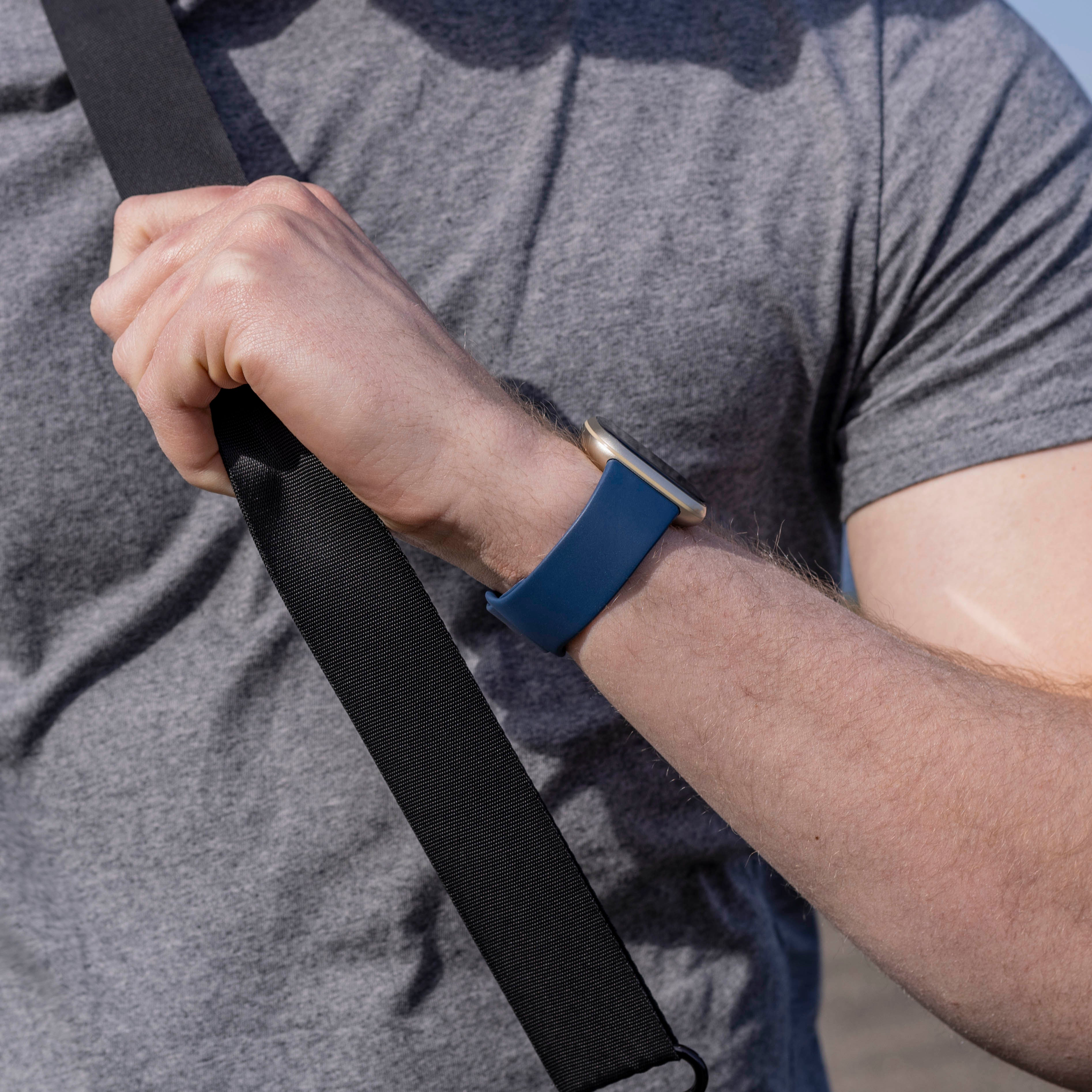 Der Arm einer Person mit einer blauen Uhr. Ein schwarzer Riemen kreuzt den Körper. Sie tragen ein graues Hemd, der Hintergrund ist blau.