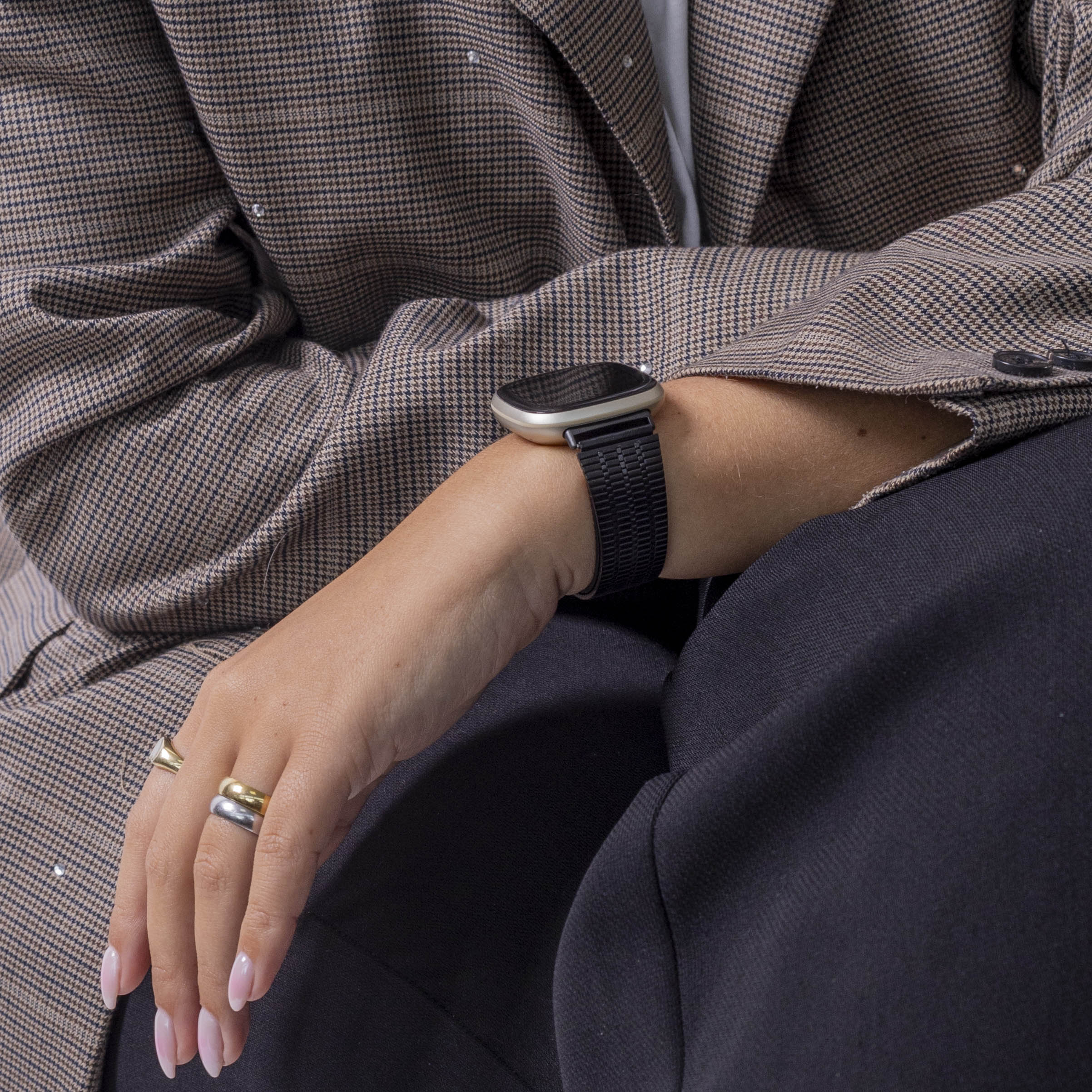 Eine Person trägt einen braunen Blazer mit Karomuster und eine schwarze Hose. Am Handgelenk ist eine Uhr. Die Hand hat Ringe.