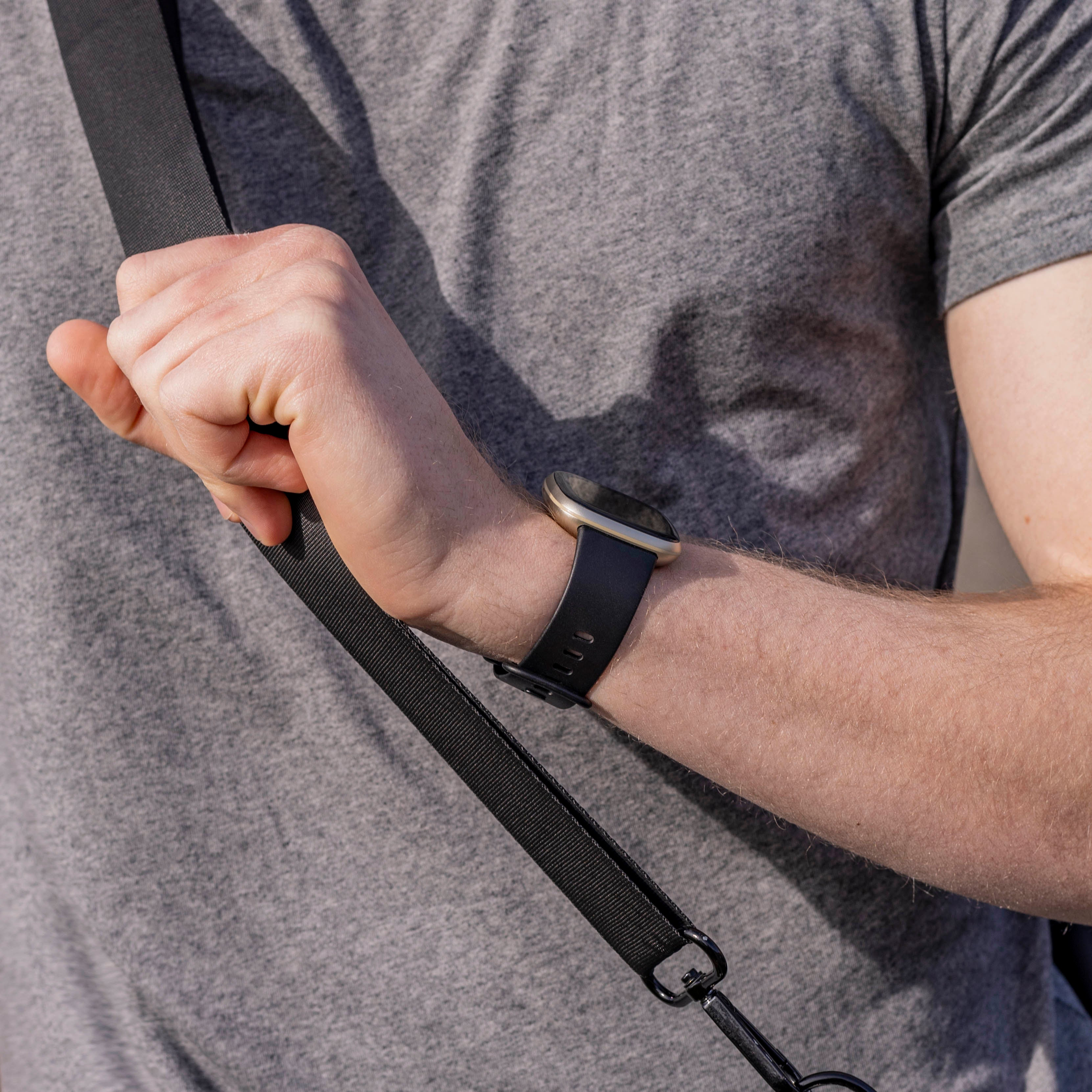 Ein Arm mit Uhr, der einen Riemen hält. Die Person trägt ein graues Hemd, der Riemen ist schwarz.