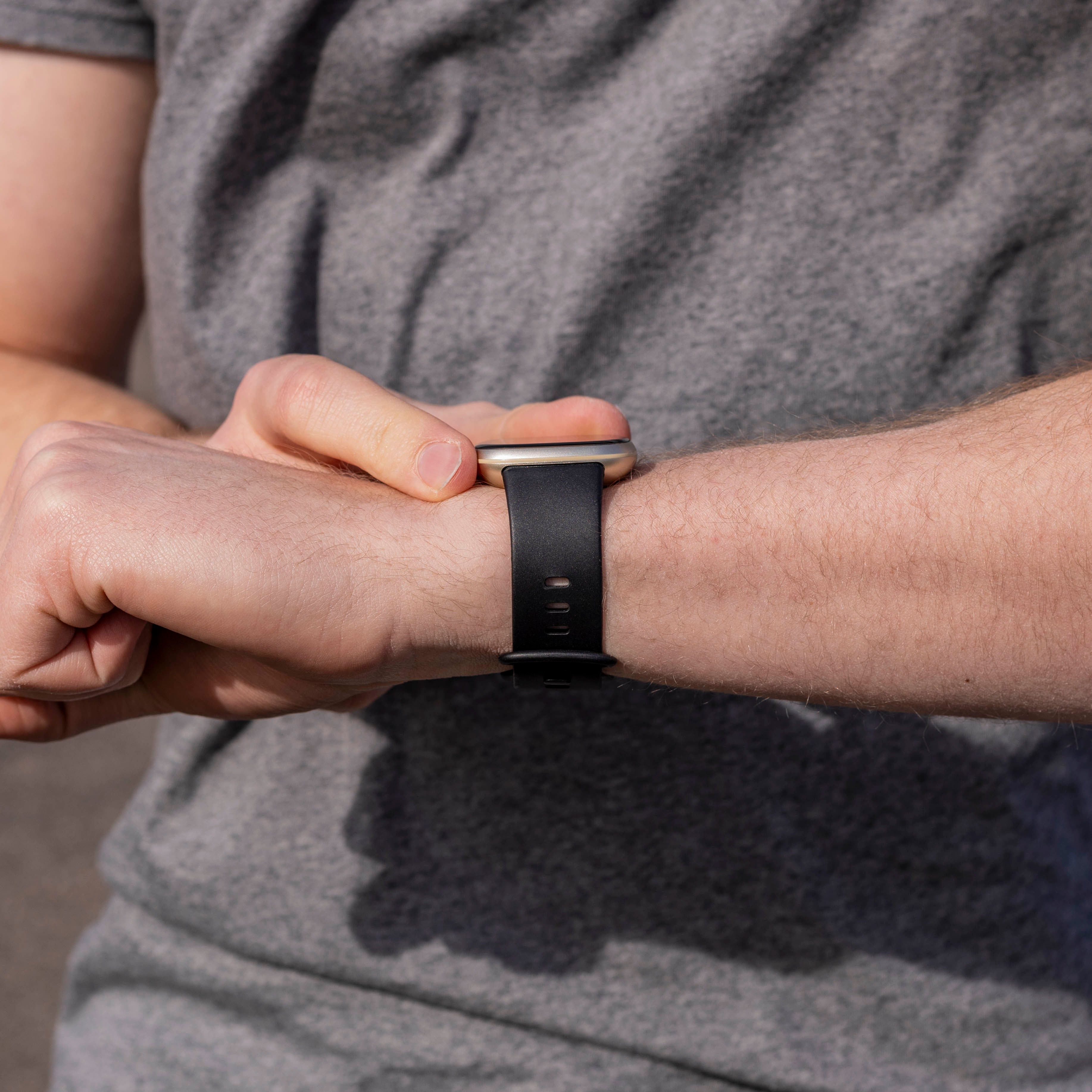 Person im grauen Hemd überprüft eine Smartwatch am linken Handgelenk. Die Uhr hat ein schwarzes Armband.