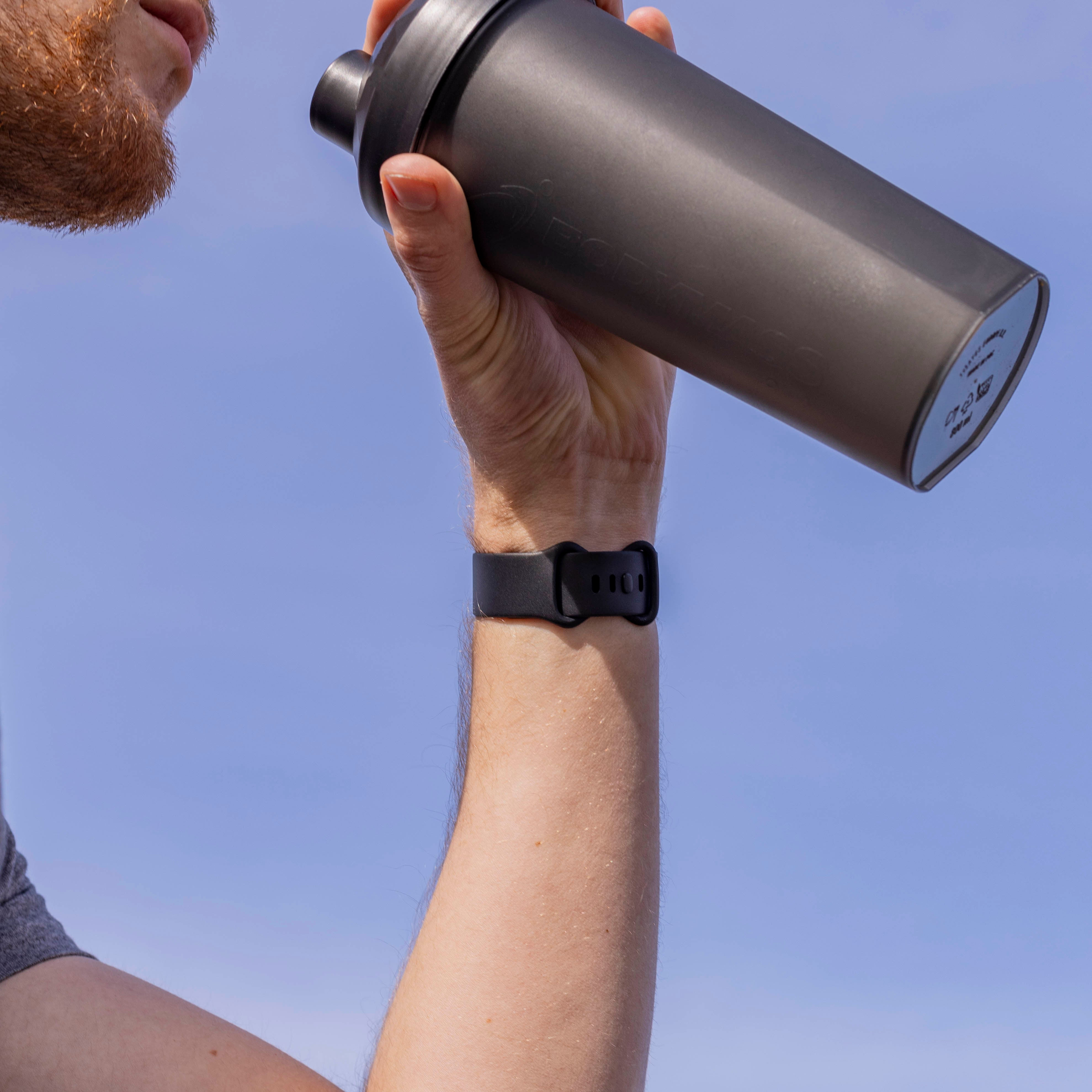 Eine Person trinkt aus einem schwarzen Shaker vor blauem Himmel. Eine schwarze Uhr ist am Handgelenk. Roter Bart ist sichtbar.