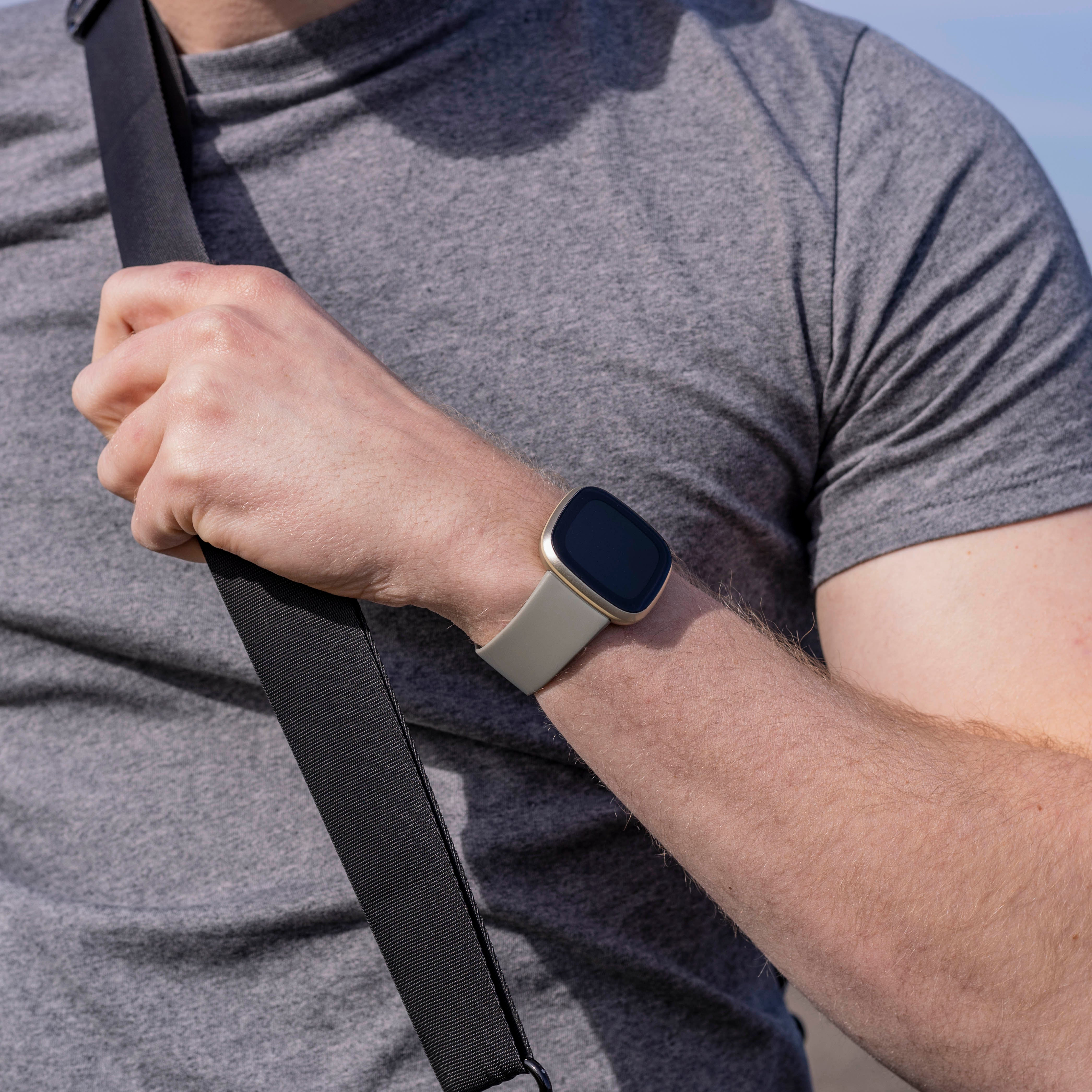 Eine Person trägt ein graues T-Shirt und eine Smartwatch mit hellgrauem Armband. Ein schwarzer Riemen ist ebenfalls sichtbar.