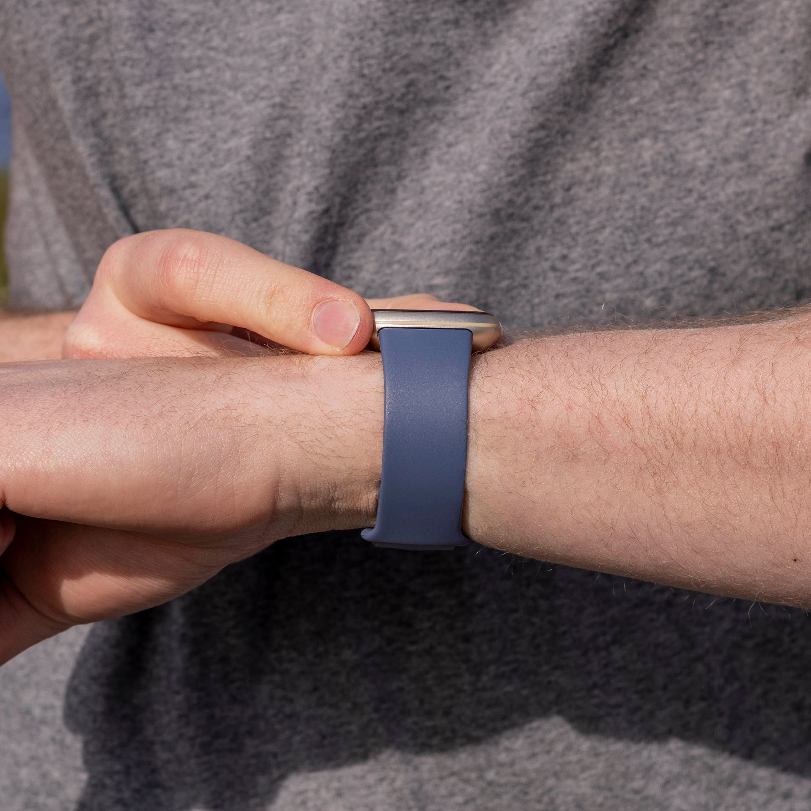 Eine Person im grauen Hemd justiert eine Smartwatch mit blauem Armband am Handgelenk. Haut ist sichtbar.