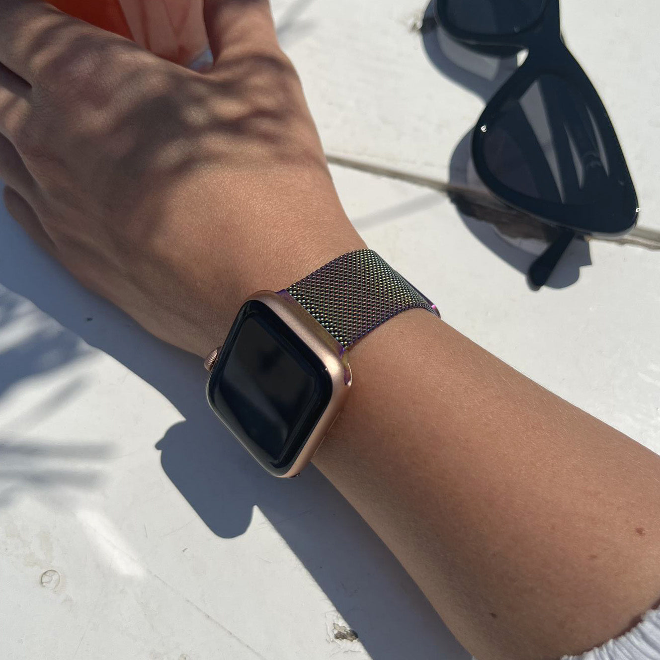 Arm mit goldener Uhr und Metallarmband. Sonnenbrille auf weißem Tisch. Die Szene ist im Freien.