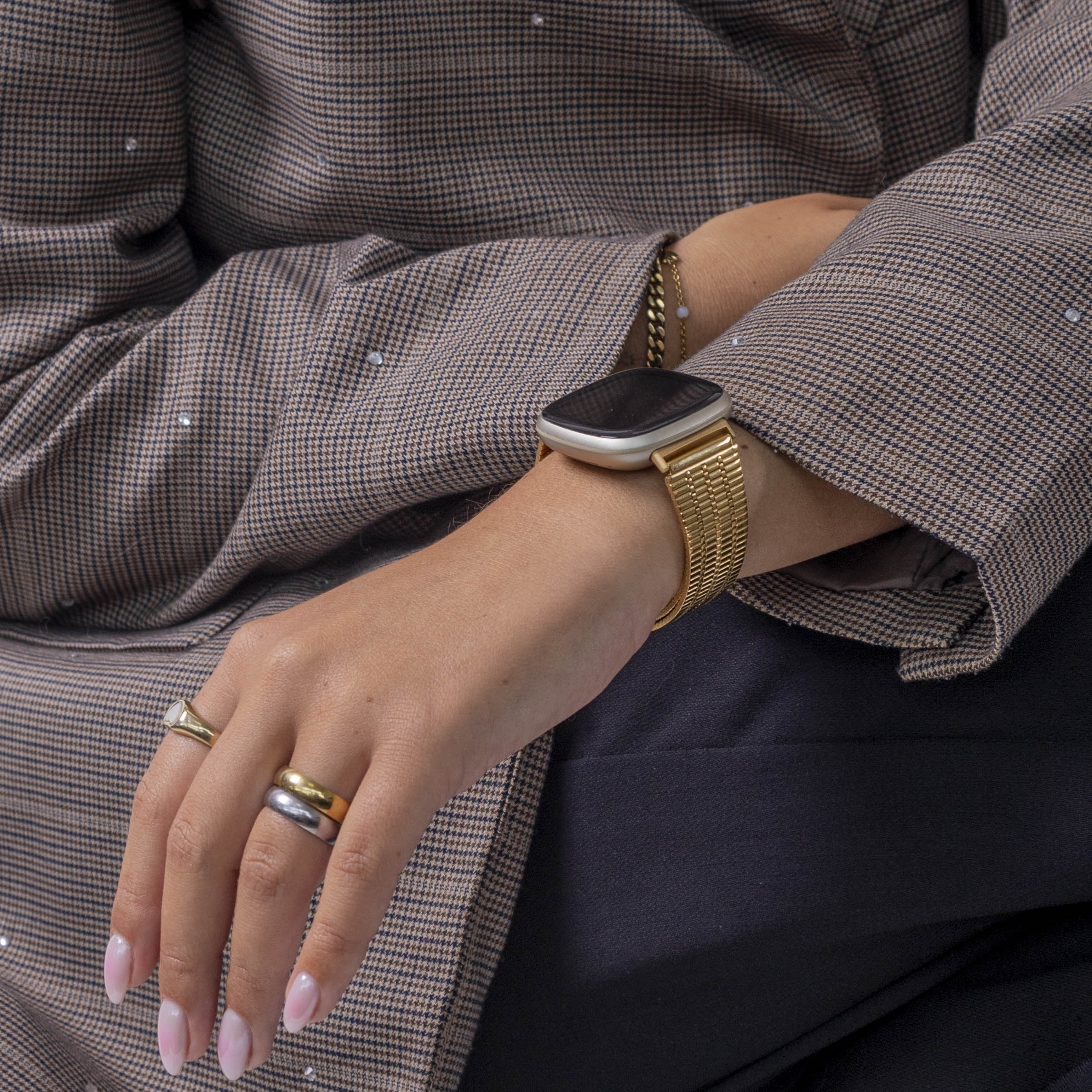 Arm einer Person mit Uhr und Ringen. Sie trägt eine braun gemusterte Jacke und dunkle Hose. Die Uhr hat ein goldenes Band.