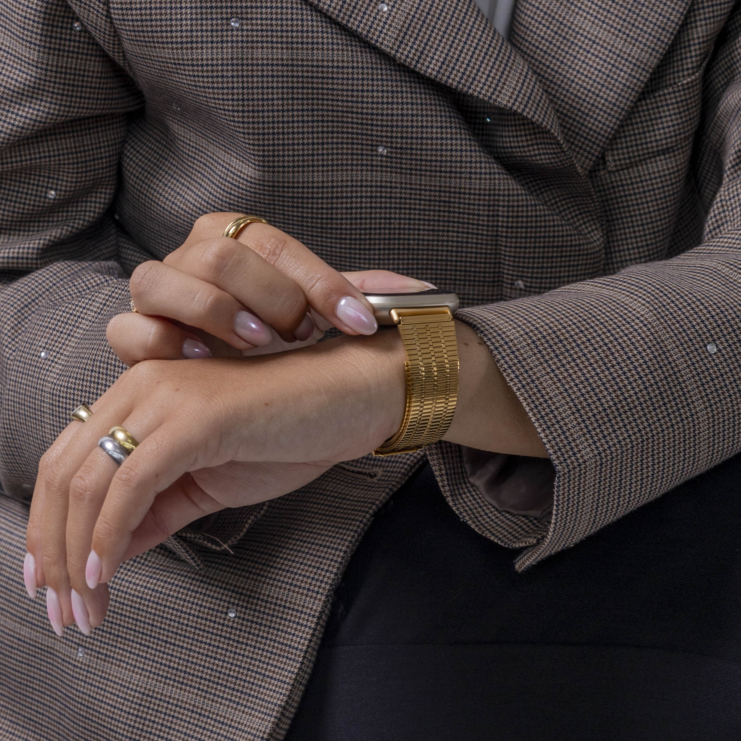 Eine Person in einem braunen Blazer mit Karomuster betrachtet eine goldene Uhr. Sie hat Ringe an den Fingern und eine schwarze Hose.