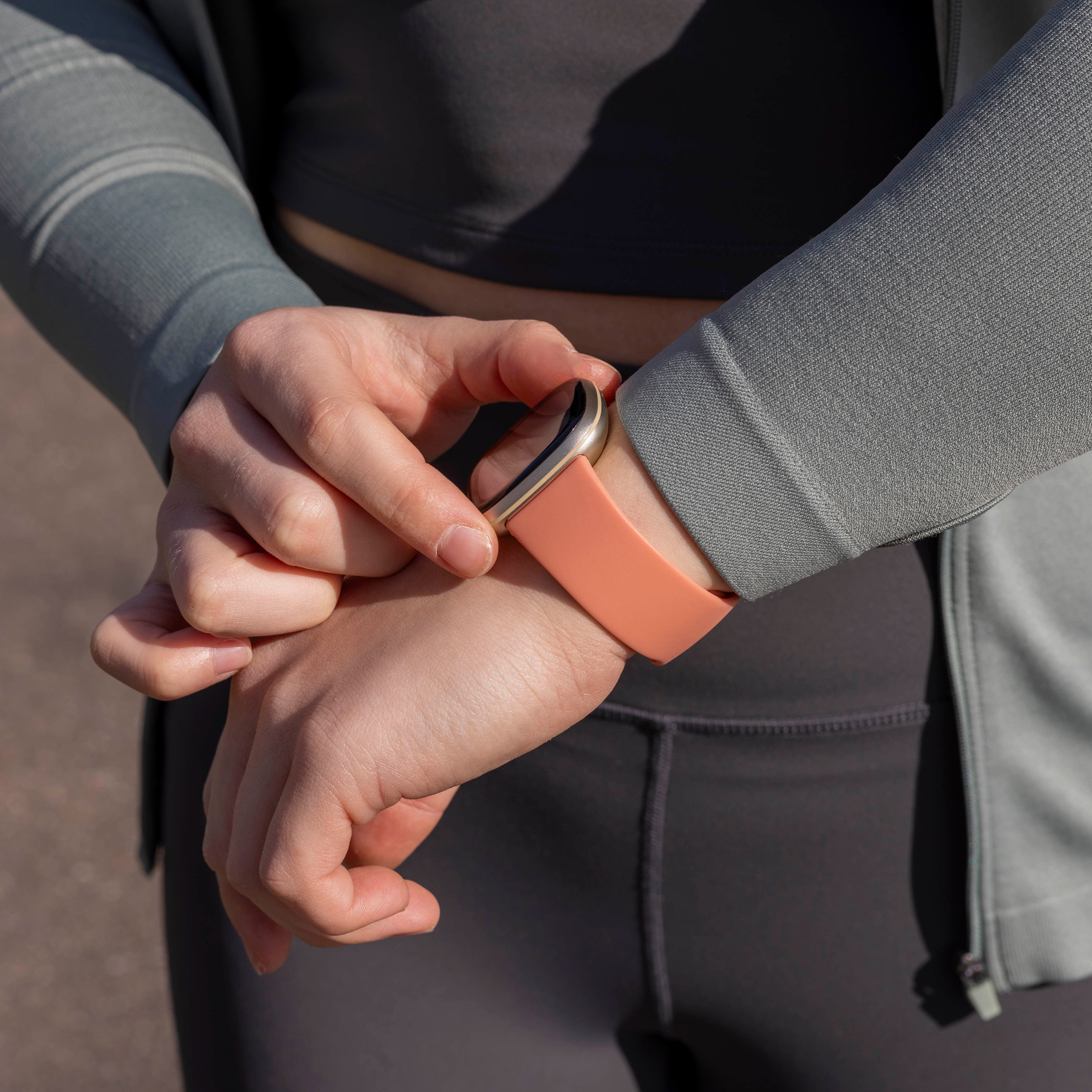 Person trägt eine Smartwatch mit einem pfirsichfarbenen Armband. Die Person trägt auch dunkle Sportkleidung und eine graue Jacke.
