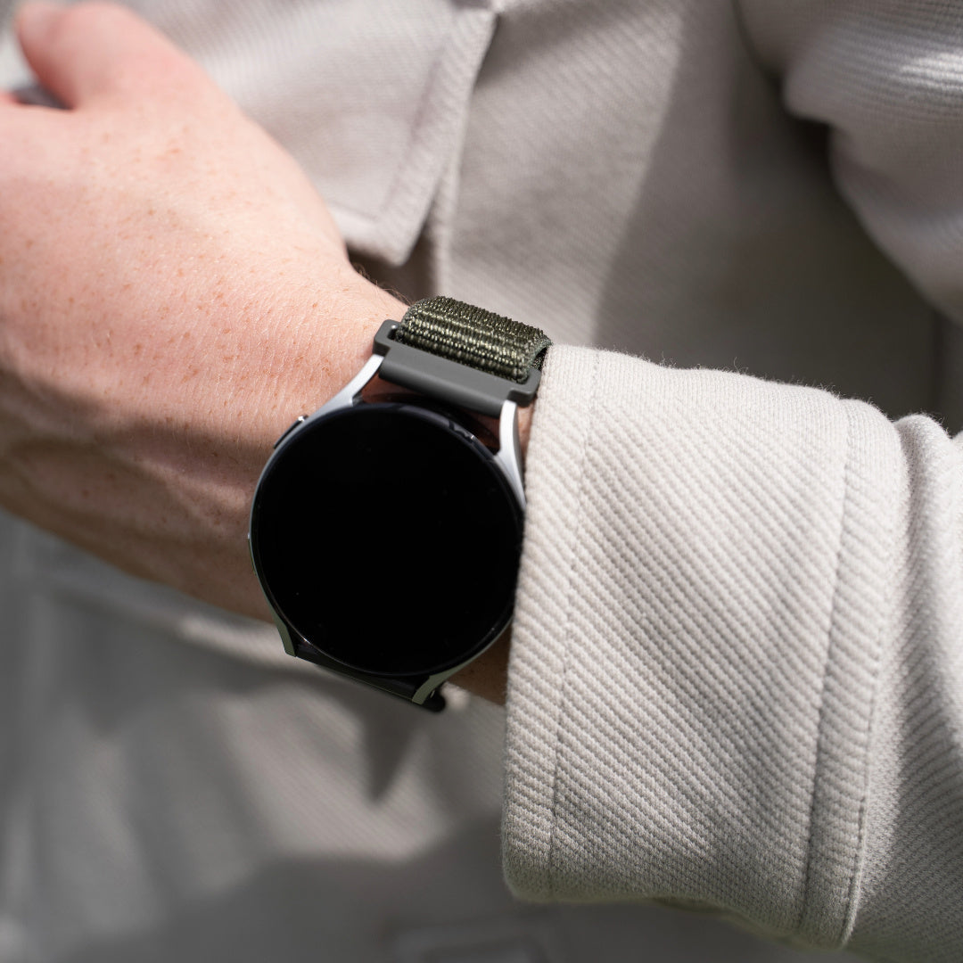 Handgelenk mit Smartwatch. Die Uhr hat einen schwarzen Bildschirm und ein grünes Stoffarmband. Der Arm ist mit einer hellen Jacke bedeckt.