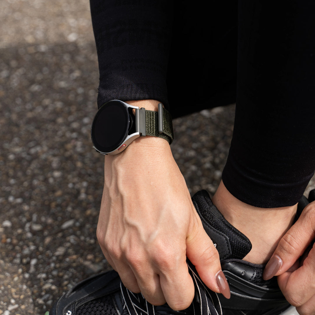 Person passt Laufschuh an, trägt Smartwatch mit grünem Armband. Asphalt Hintergrund, schwarze Hose, helle Haut und schwarzer Sneaker.