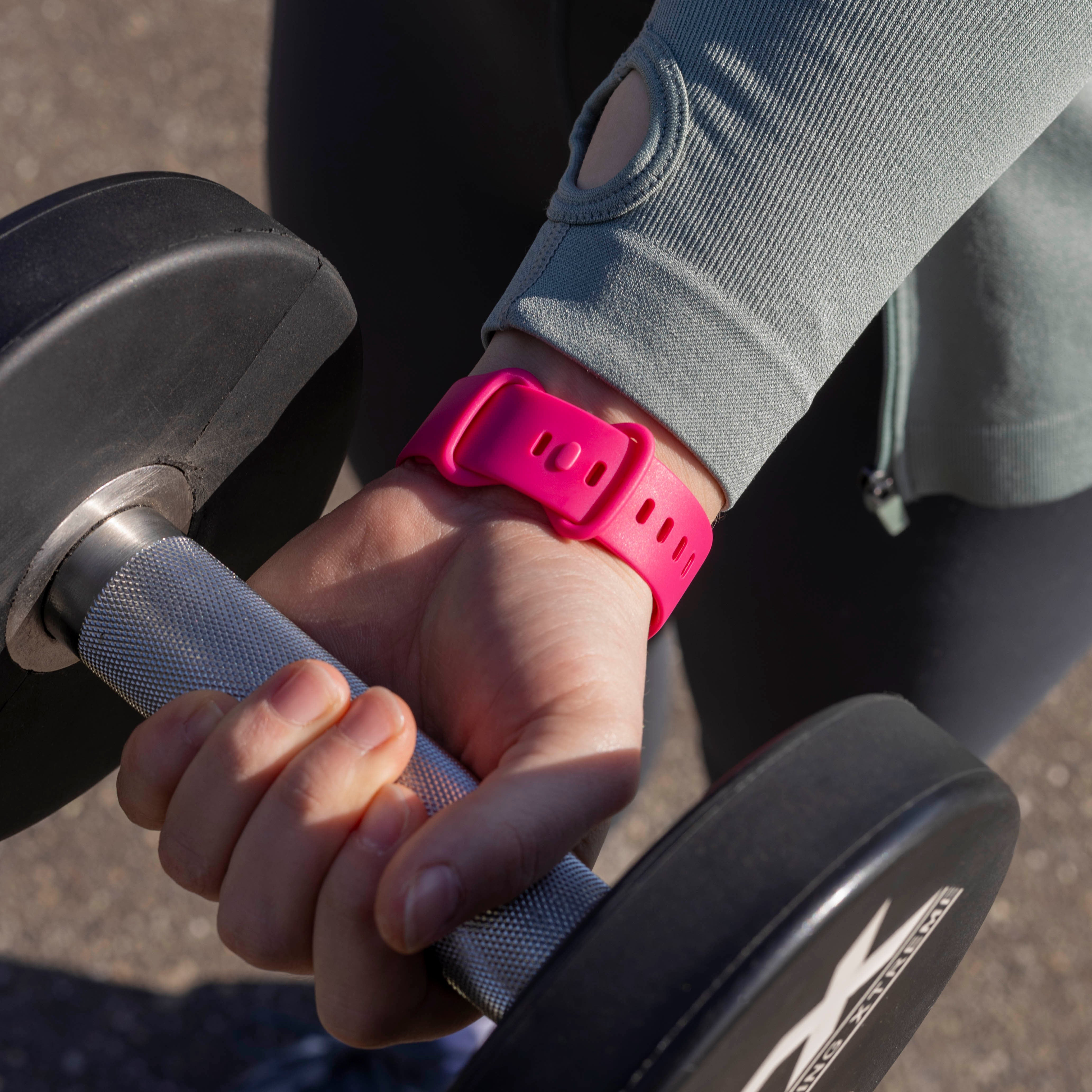 Die Hand einer Person hält eine Hantel. Eine pinkfarbene Smartwatch befindet sich am Handgelenk. Die Person trägt ein graues Hemd.