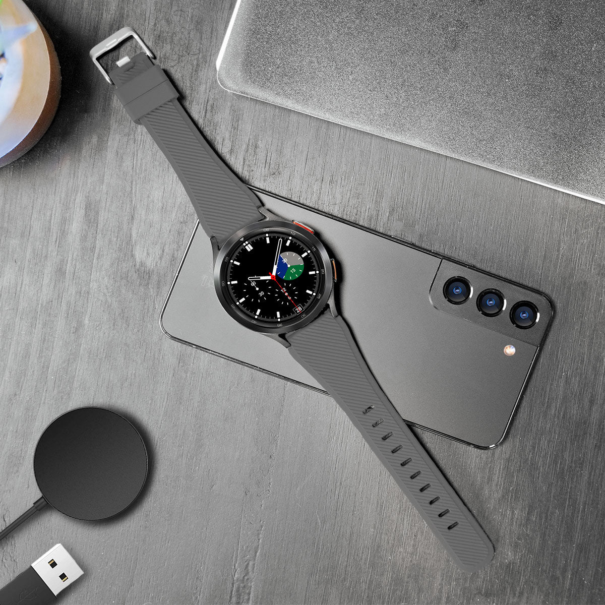 Eine graue Smartwatch mit schwarzem Zifferblatt und grauem Armband liegt auf einem grauen Smartphone. Außerdem sind ein Ladepad und ein Laptop sichtbar.