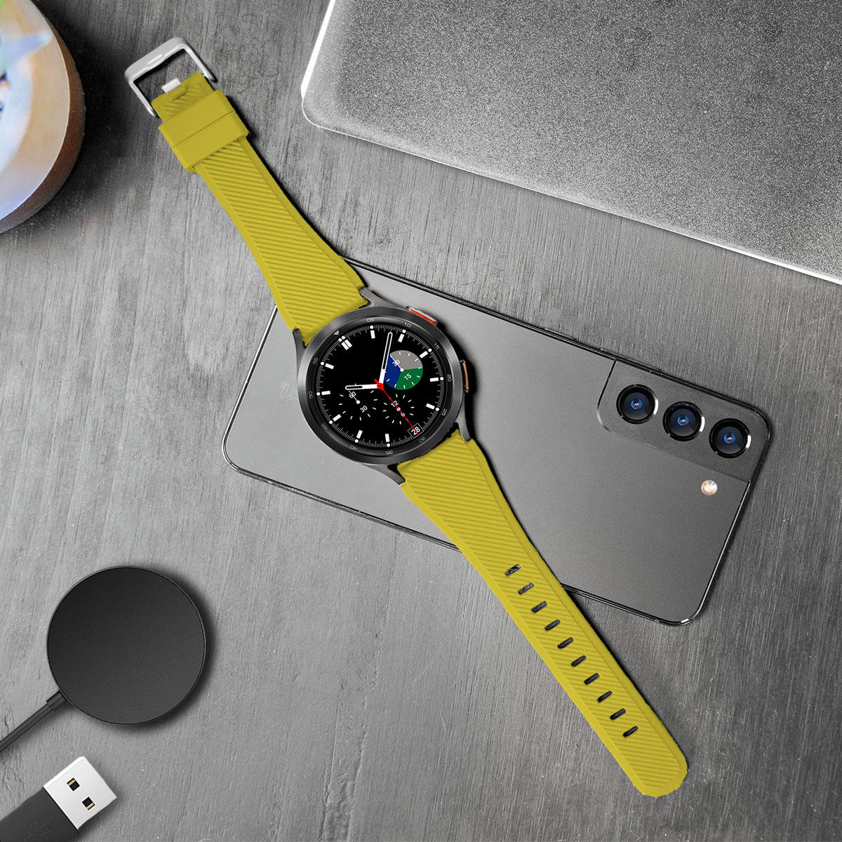 Eine Smartwatch mit gelbem Armband liegt auf einem Telefon, einem Laptop und einem Ladegerät.