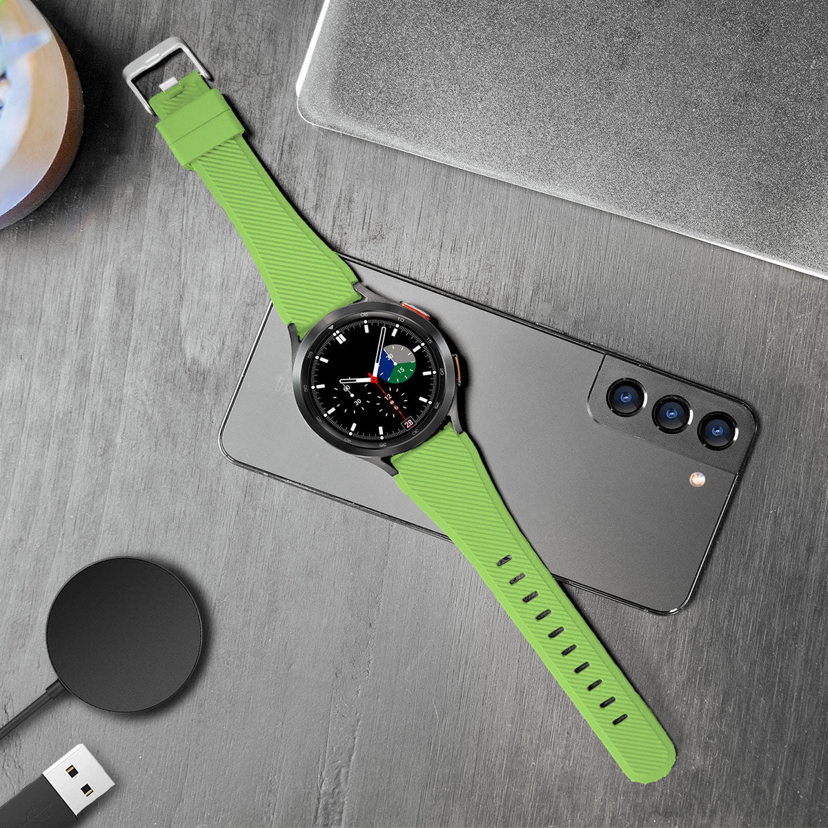 Eine grüne Smartwatch mit schwarzem Display, ein Smartphone und ein Ladepad liegen auf einer grauen Oberfläche. Im Hintergrund ist ein Laptop.