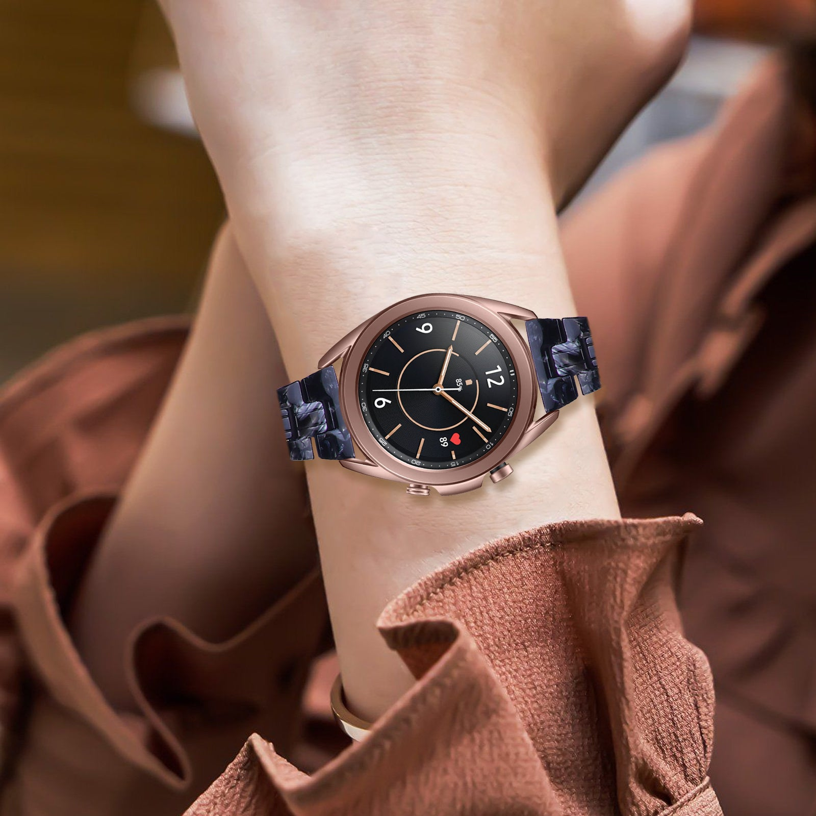 Eine roségoldene Smartwatch mit schwarzem Zifferblatt und blauem Marmorarmband befindet sich an einem Handgelenk. Die Person trägt ein braunes Oberteil.