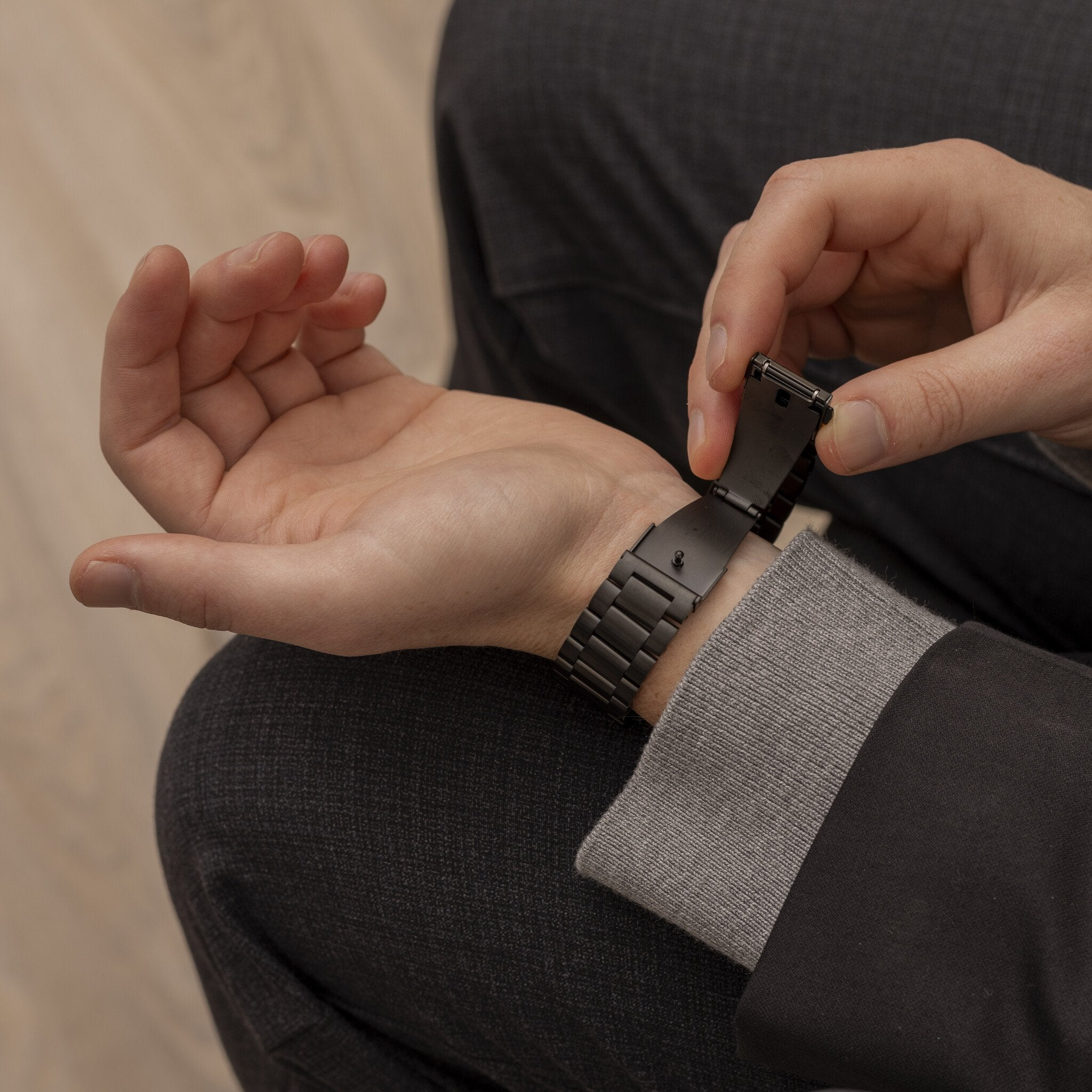 Eine Person befestigt eine schwarze Uhr an ihrem Handgelenk. Die Person trägt einen grauen Pullover und eine schwarze Jacke.