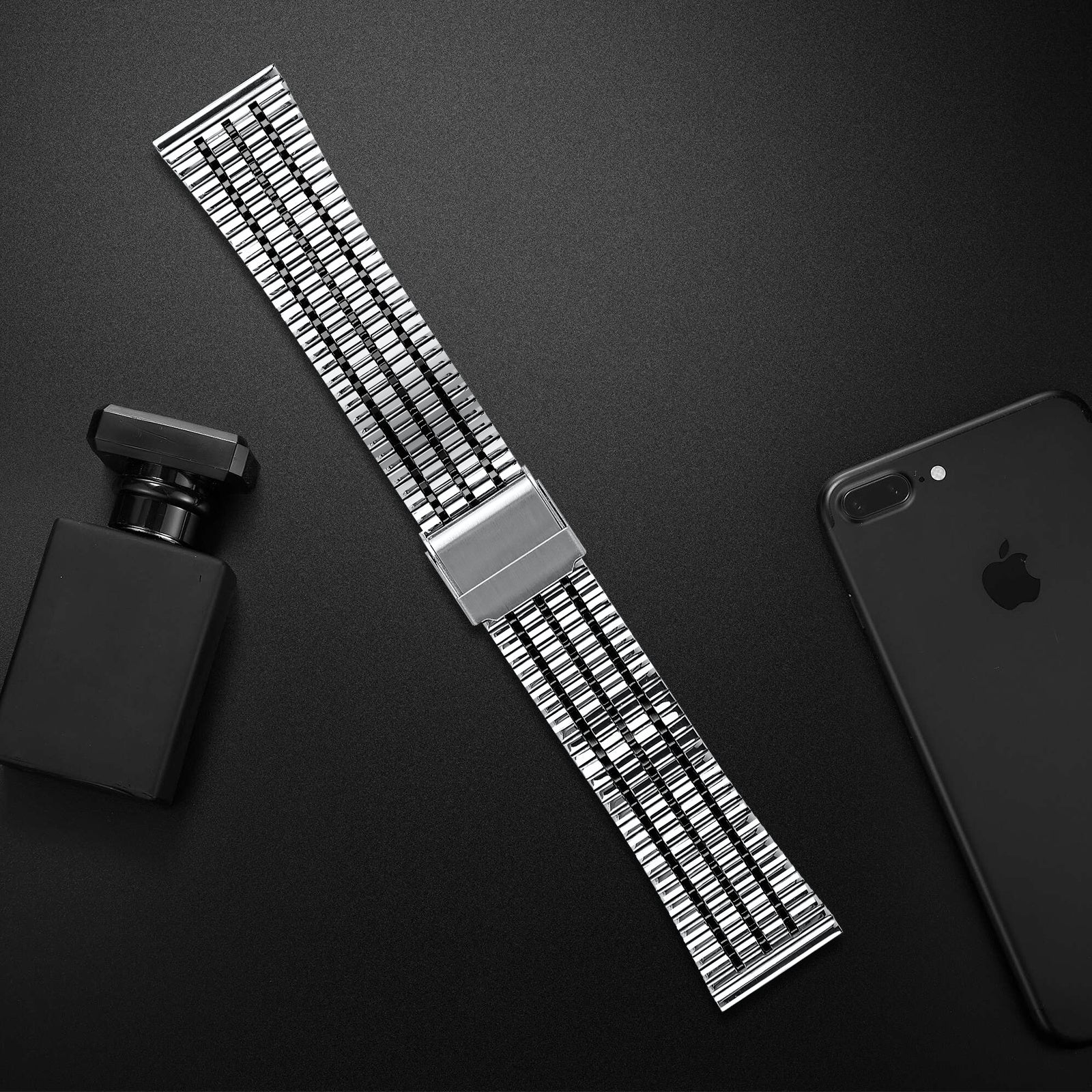Ein Metall-Uhrenarmband, eine Parfümflasche und ein schwarzes Smartphone liegen auf einer dunklen Oberfläche. Das Armband ist zentriert, Objekte sind drumherum.
