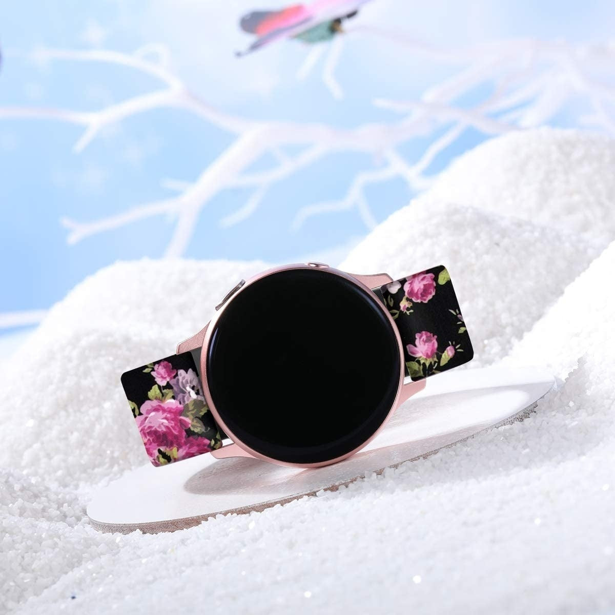 Eine roségoldene Smartwatch mit schwarzem Bildschirm und floralen Armbändern liegt auf einer weißen Oberfläche, umgeben von Kunstschnee. Im Hintergrund ist ein blauer Himmel zu sehen.