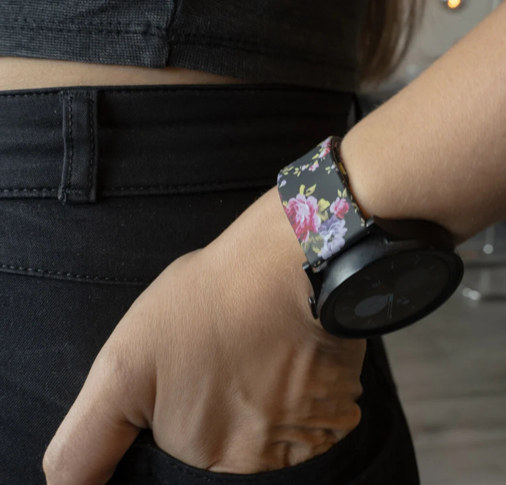 Arm einer Person mit einer schwarzen Smartwatch mit floralem Band, die auf einer schwarzen Jeans ruht. Die Hand der Person ist in der Tasche.