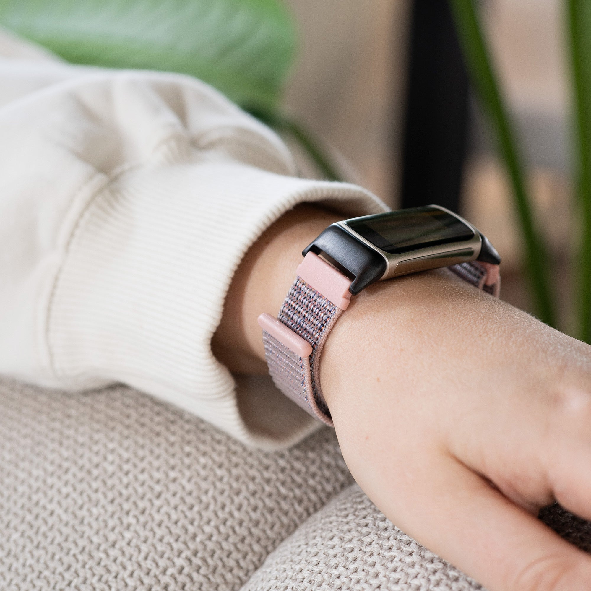 Das Handgelenk einer Person mit einer Smartwatch. Die Uhr hat einen schwarzen Bildschirm und ein rosa-graues Armband. Die Person trägt einen beigefarbenen Pullover.