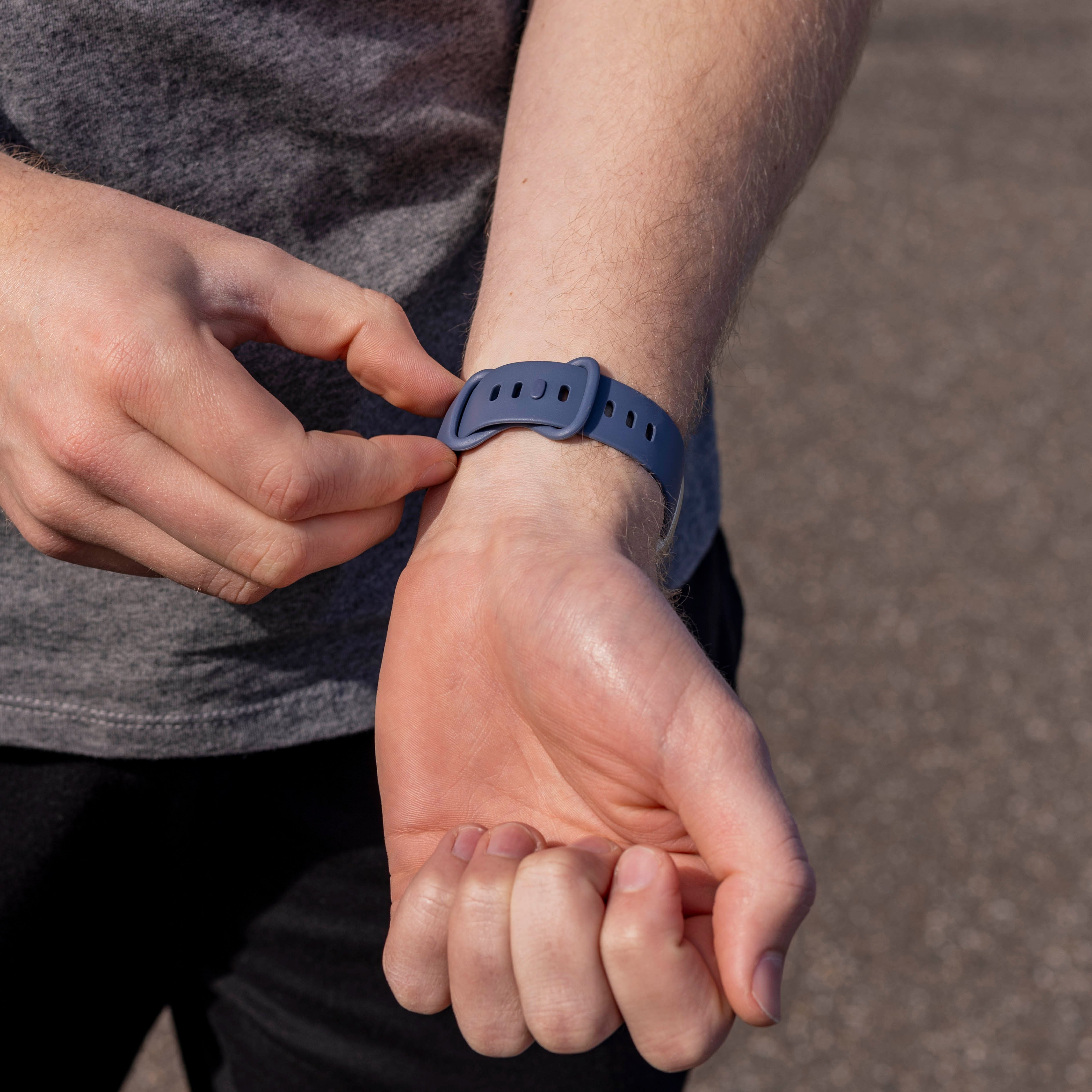 Eine Person stellt ein blaues Uhrenarmband an ihrem Handgelenk ein. Sie trägt ein graues Hemd und schwarze Hose. Der Hintergrund ist unscharf.