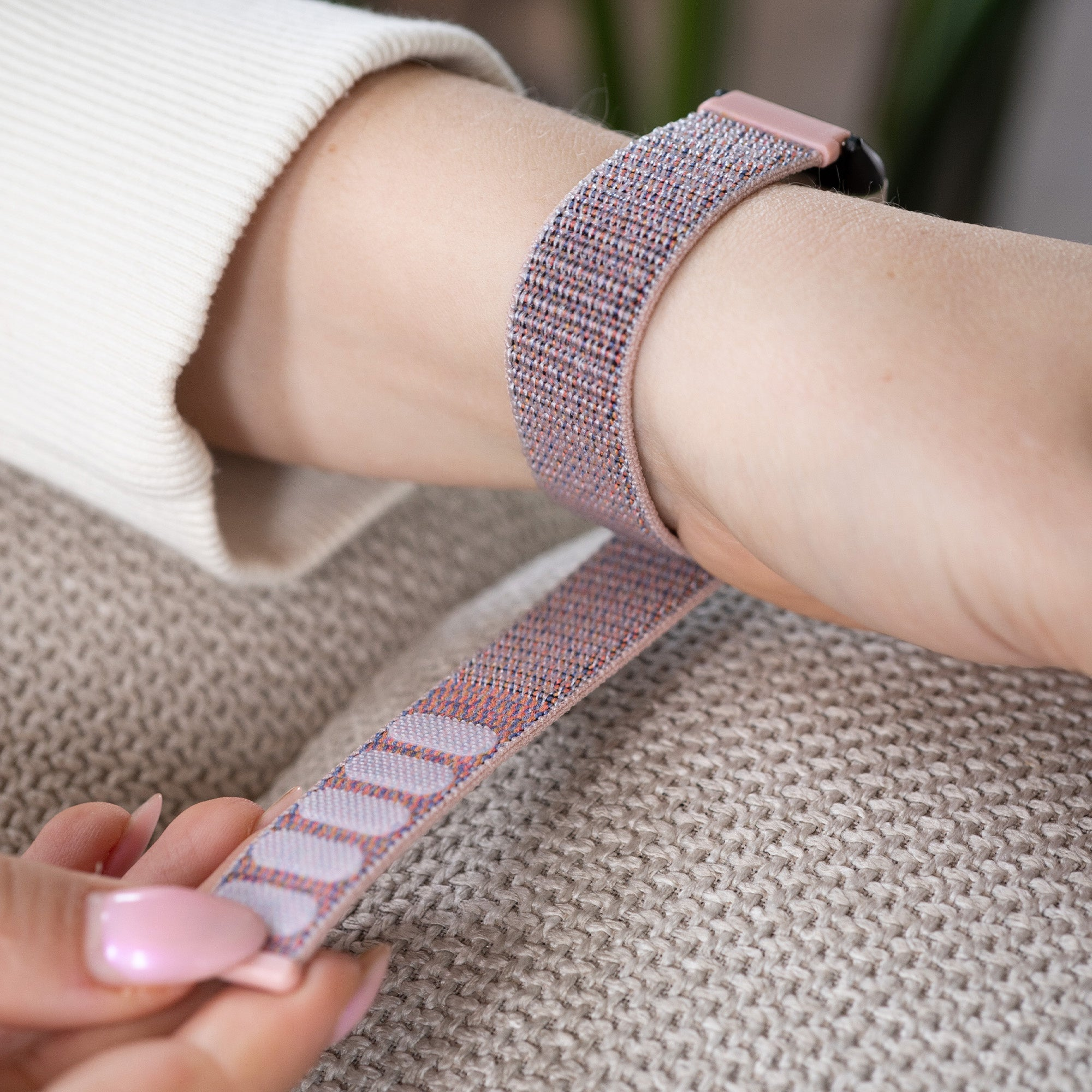 Ein Handgelenk mit einem rosafarbenen Stoffarmband wird angepasst. Die Person trägt einen cremefarbenen Pullover und sitzt auf einer beigen Oberfläche.